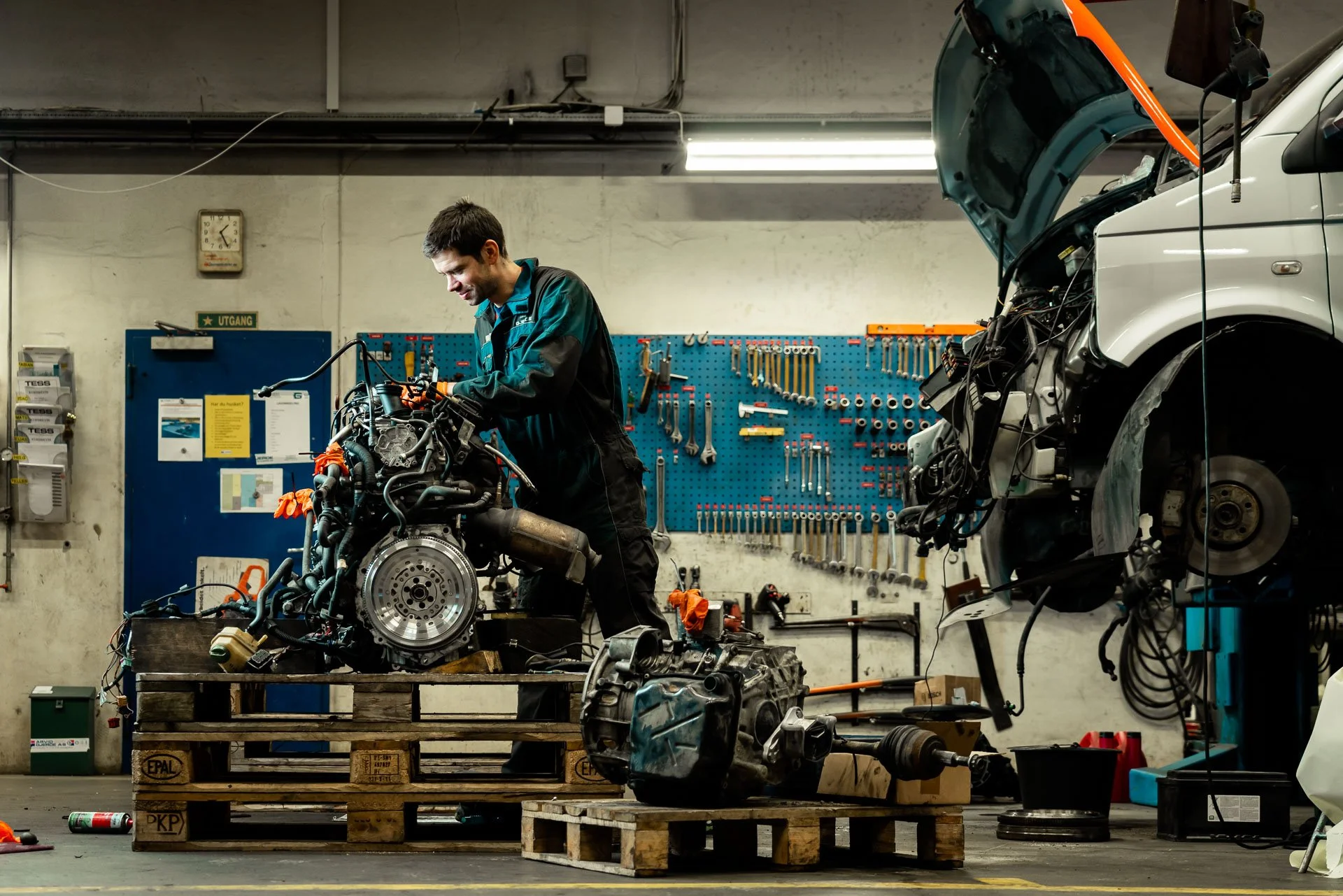 En mann som jobber med en motor i et verksted, omgitt av verktøy, en bil uten hjul, og motorer plassert på paller.