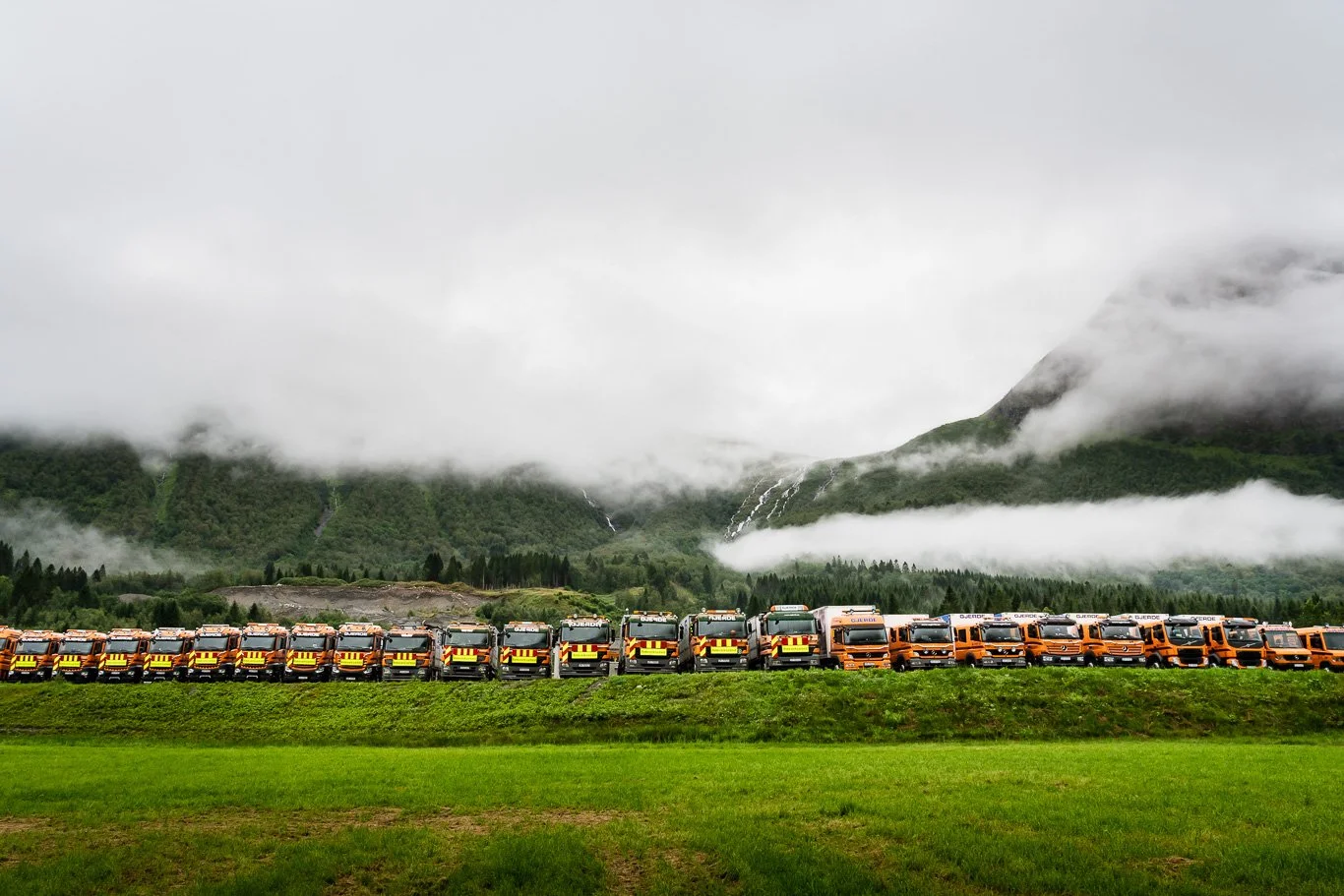 Lastebilene fra Arvid Gjerde oppstilt foran kontorbygget og verkstedet i Innfjorden i Rauma. I bakgrunnen er det fjellandskap med skog og lave skyer.