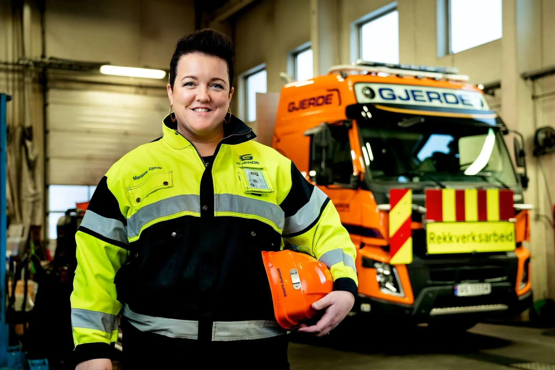 Marianne Gjerde, daglig leder i Arvid Gjerde AS, i garasjen/verkstedet. Hun har på seg arbeidstøy og holder hjelmen i hånden. I bakgrunnen står en lastebil