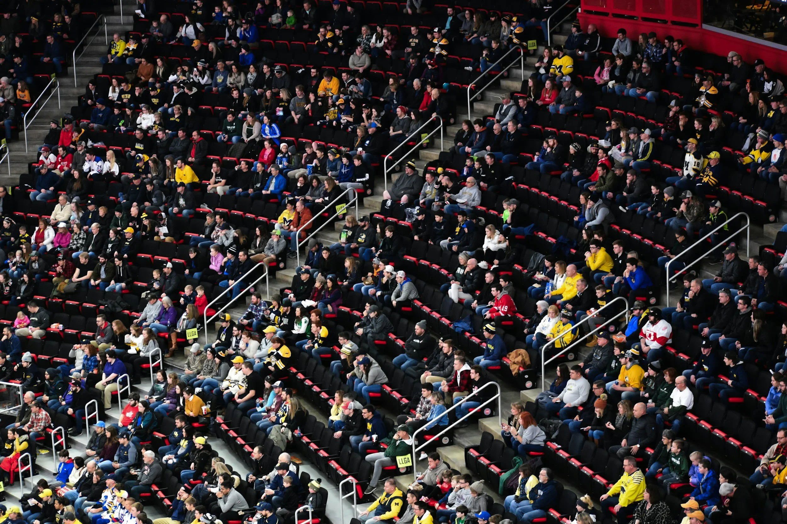 Crowd of people seated in stadium bleachers, watching an event, many wearing sports jerseys and hats.