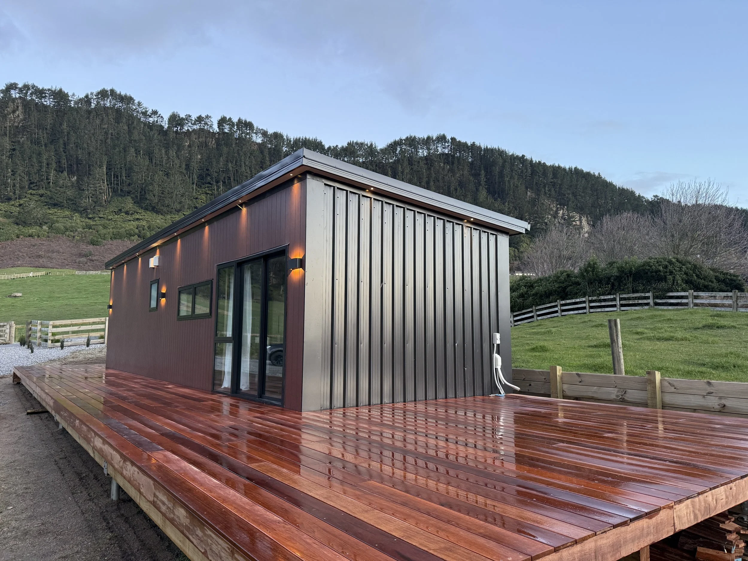 Modern tiny house with metal siding and large sliding glass door, situated on a wet wooden deck with a rural landscape in the background.