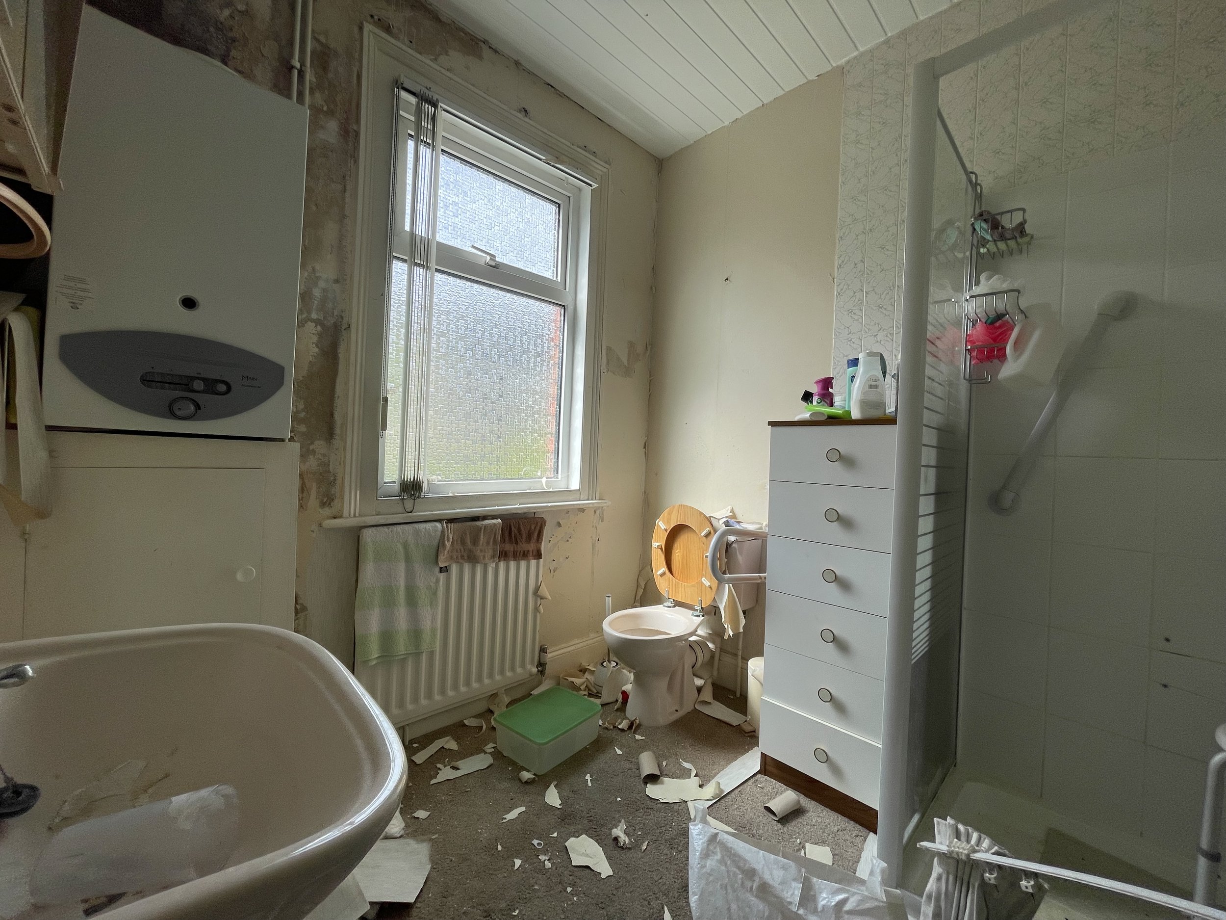 Unkempt bathroom with sink, toilet, shower, boiler, and a window.