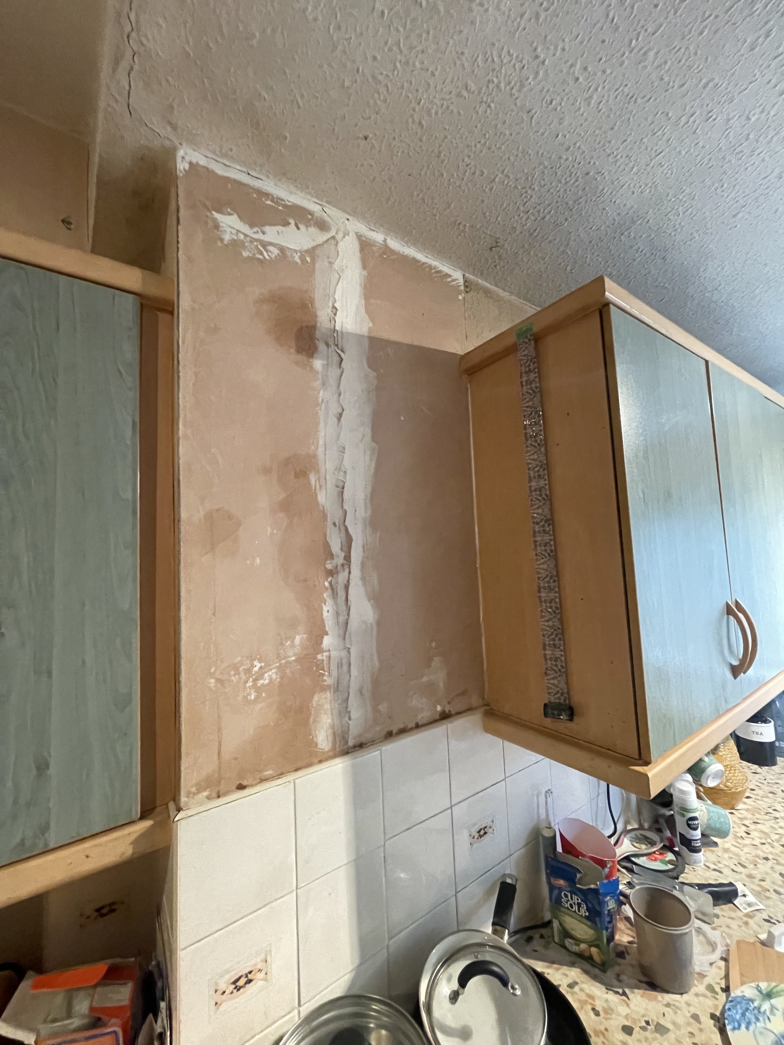 Kitchen corner with wall repair, wooden cabinets, white tiled backsplash, and countertop cluttered with cooking items.