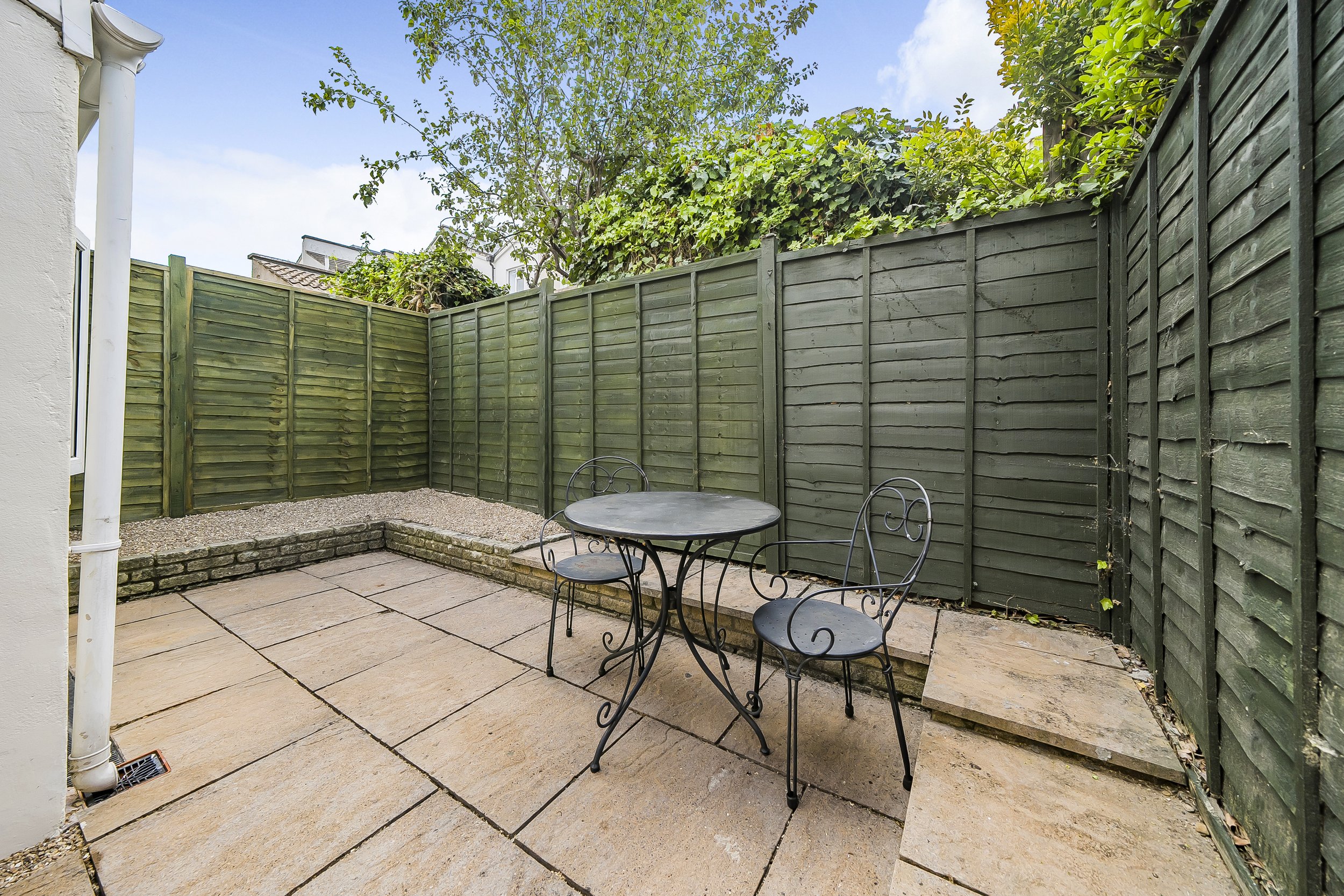 Outdoor patio with a black metal table and two chairs, surrounded by green wooden fences and greenery.
