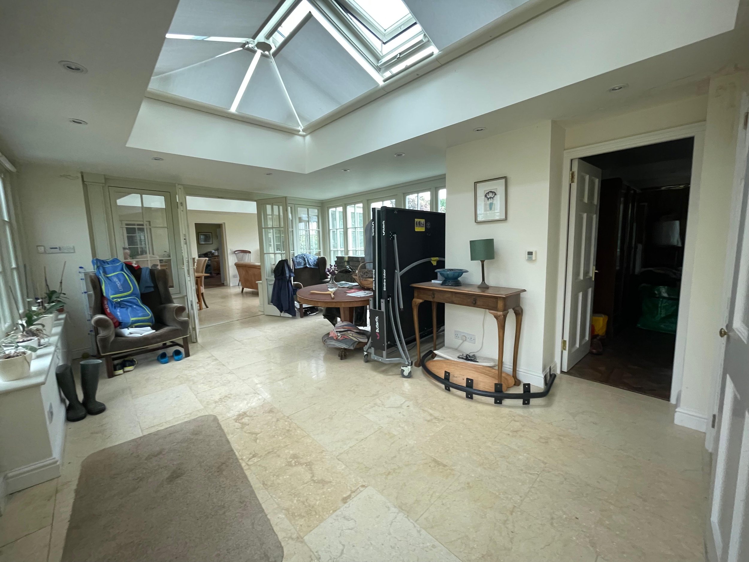 Bright conservatory interior with skylight, tile flooring, and indoor plants. There's furniture including chairs, a table, and a side table with a lamp. Two pairs of boots are visible, and a door leads to another room.