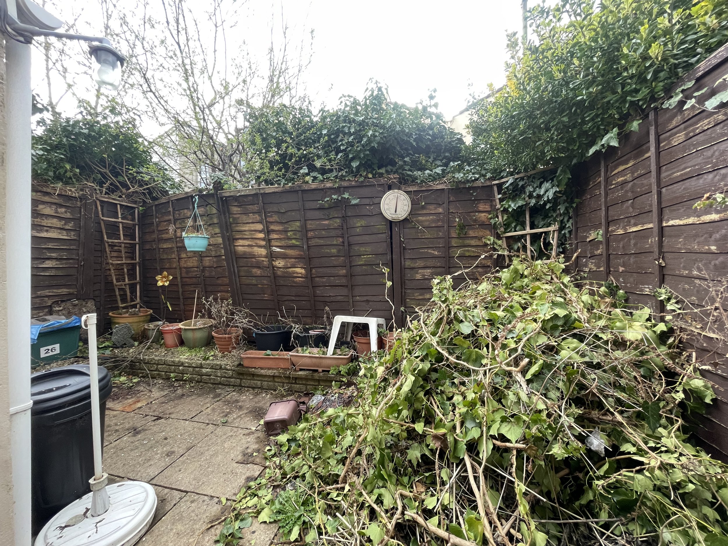 Backyard with a wooden fence, overgrown plants, hanging pots, and garden debris on the ground. A white plastic chair and a pile of green branches and leaves are visible. A wall-mounted clock and a plastic trash can are also present.