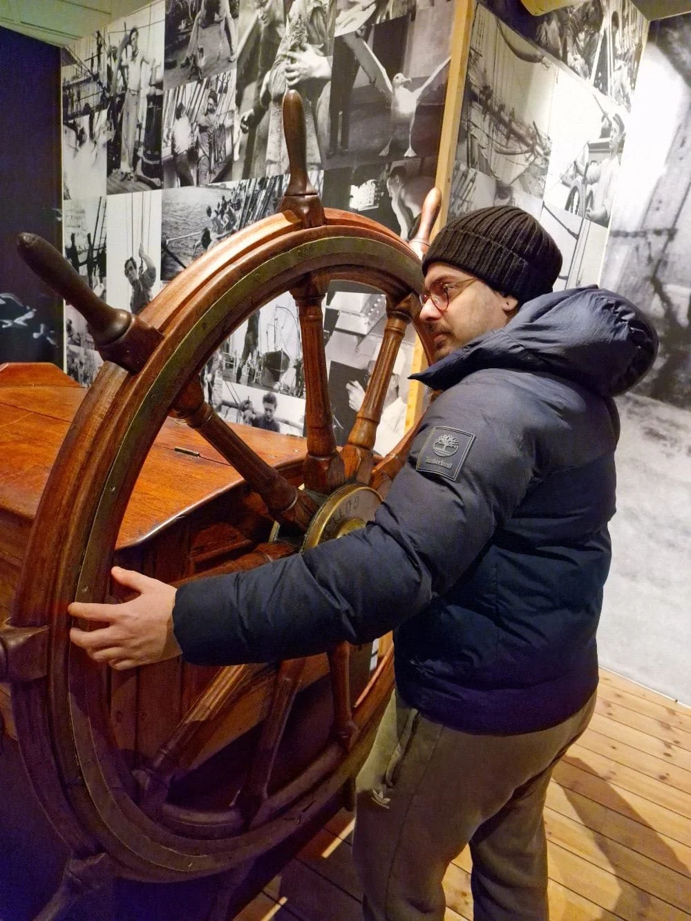 Homem de óculos, gorro preto e jaqueta acolchoada observa um leme de navio antigo em uma exposição, com paredes decoradas com fotos em preto e branco.