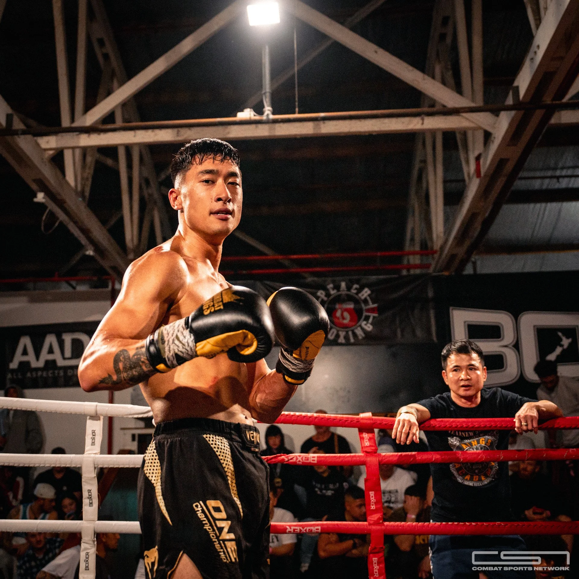 A male boxer with boxing gloves inside a boxing ring, with a trainer or coach leaning on the ring ropes outside. The background shows spectators and a gym setting.