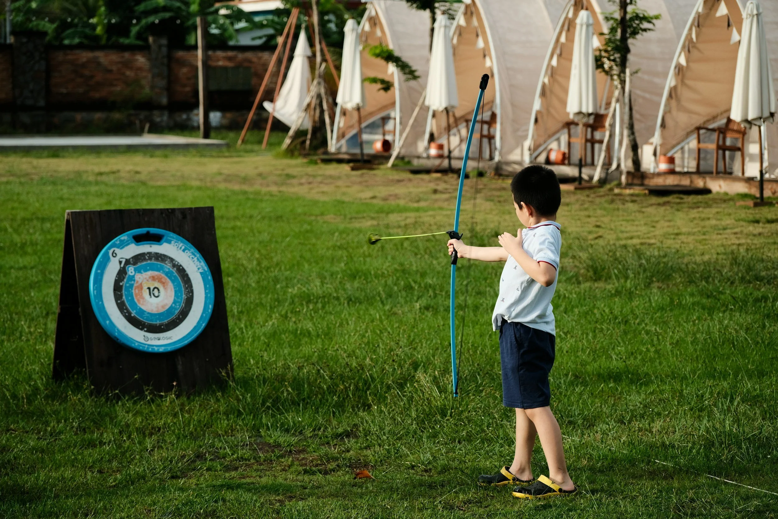 Child practicing archery with a blue bow aiming at a target on a grass field.