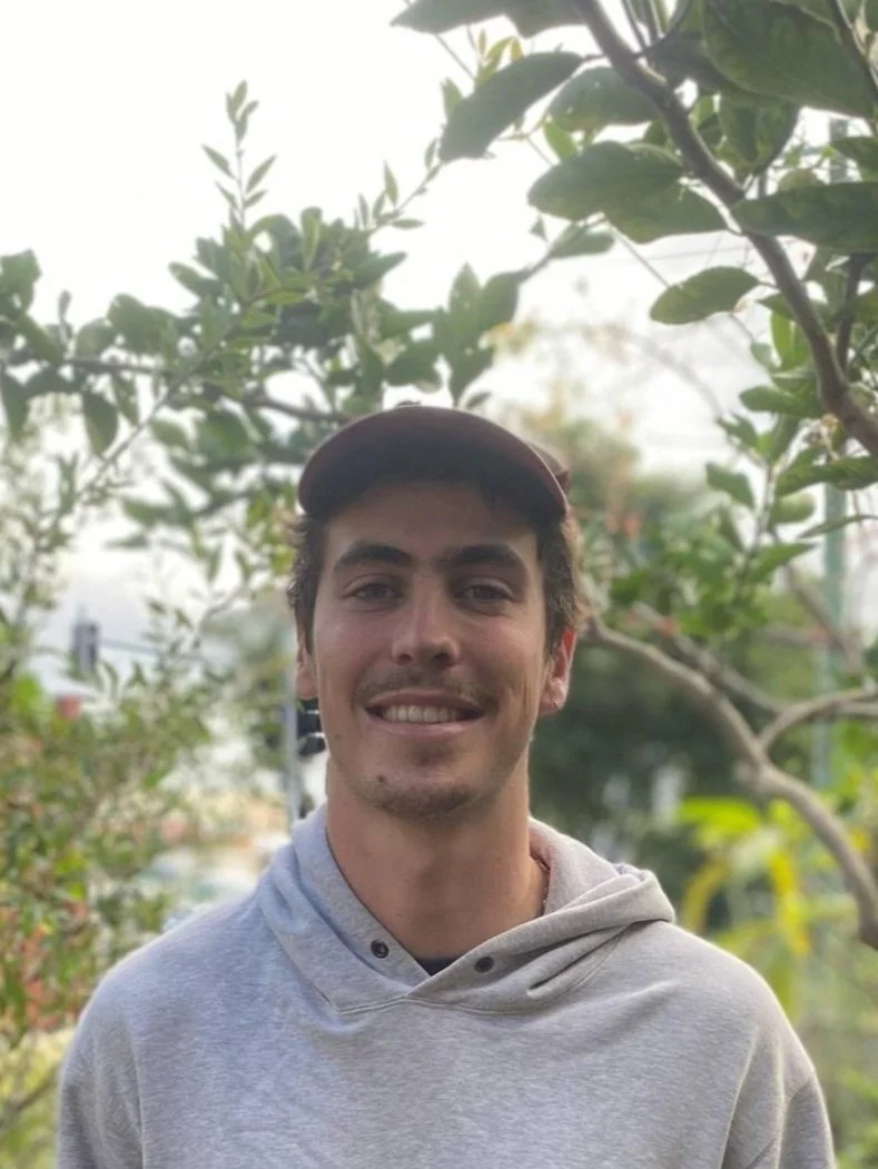 Man wearing a gray hoodie and a cap smiling in a garden setting.