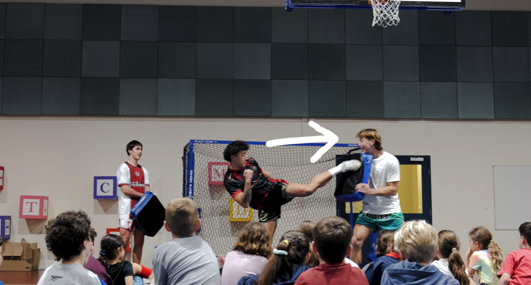 Two young men demonstrating a martial arts or kickboxing move in front of an audience of children. One is executing a high roundhouse kick towards a padded target held by the other. The scene appears to be part of a training or educational session in a gym or dojo.