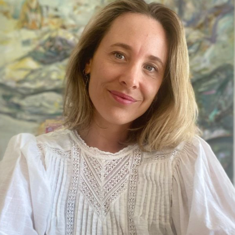 A woman with shoulder-length blonde hair and blue eyes smiling, wearing a white embroidered blouse, with a colorful abstract painting in the background.