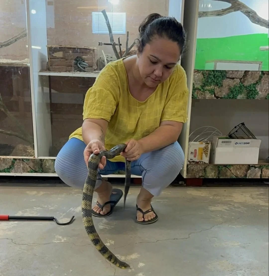 Woman in yellow shirt and flip-flops holding a snake inside a zoo or pet enclosure.