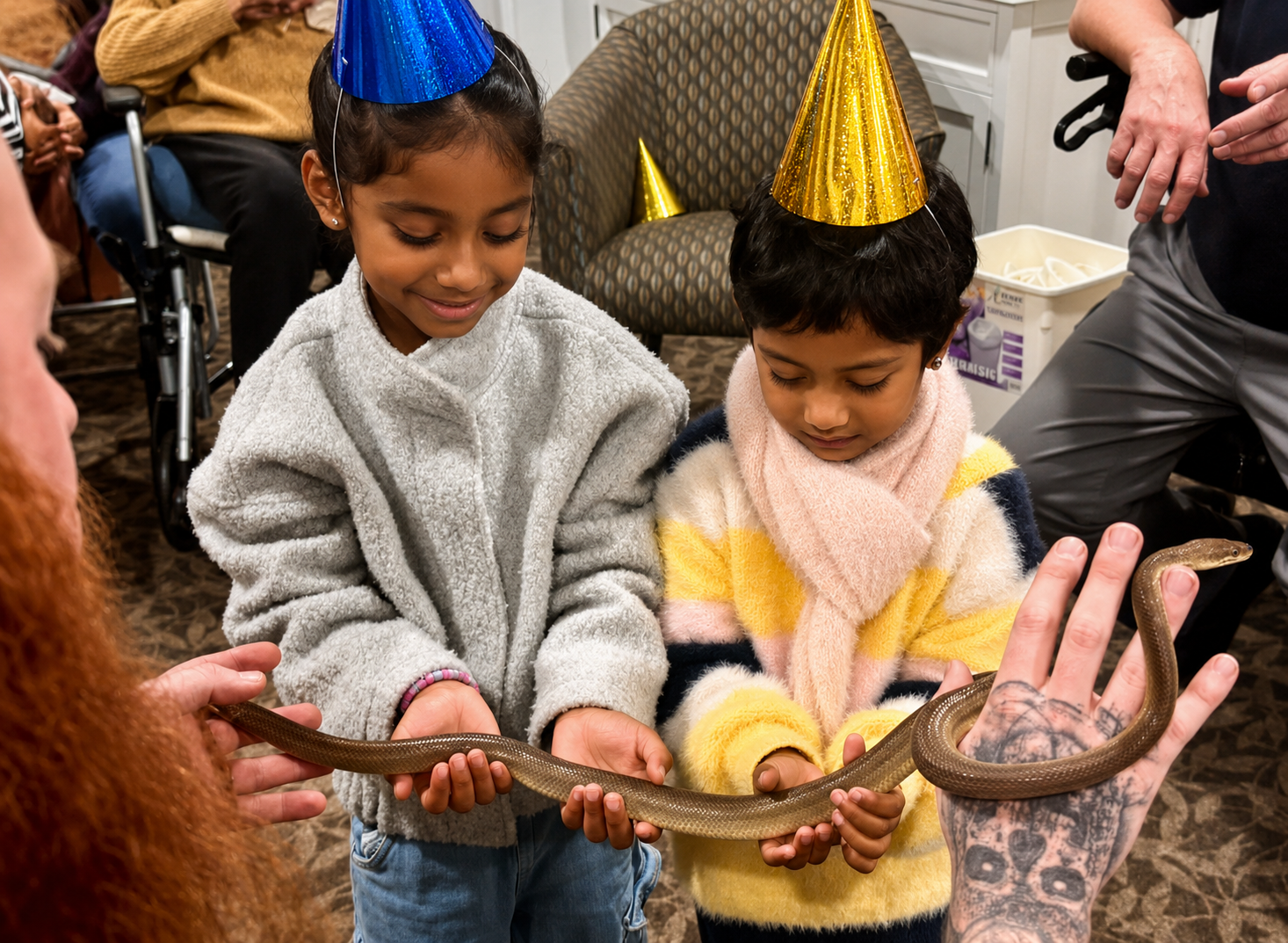 Two young girls wearing party hats holding a snake with the help of two adults at a birthday celebration.