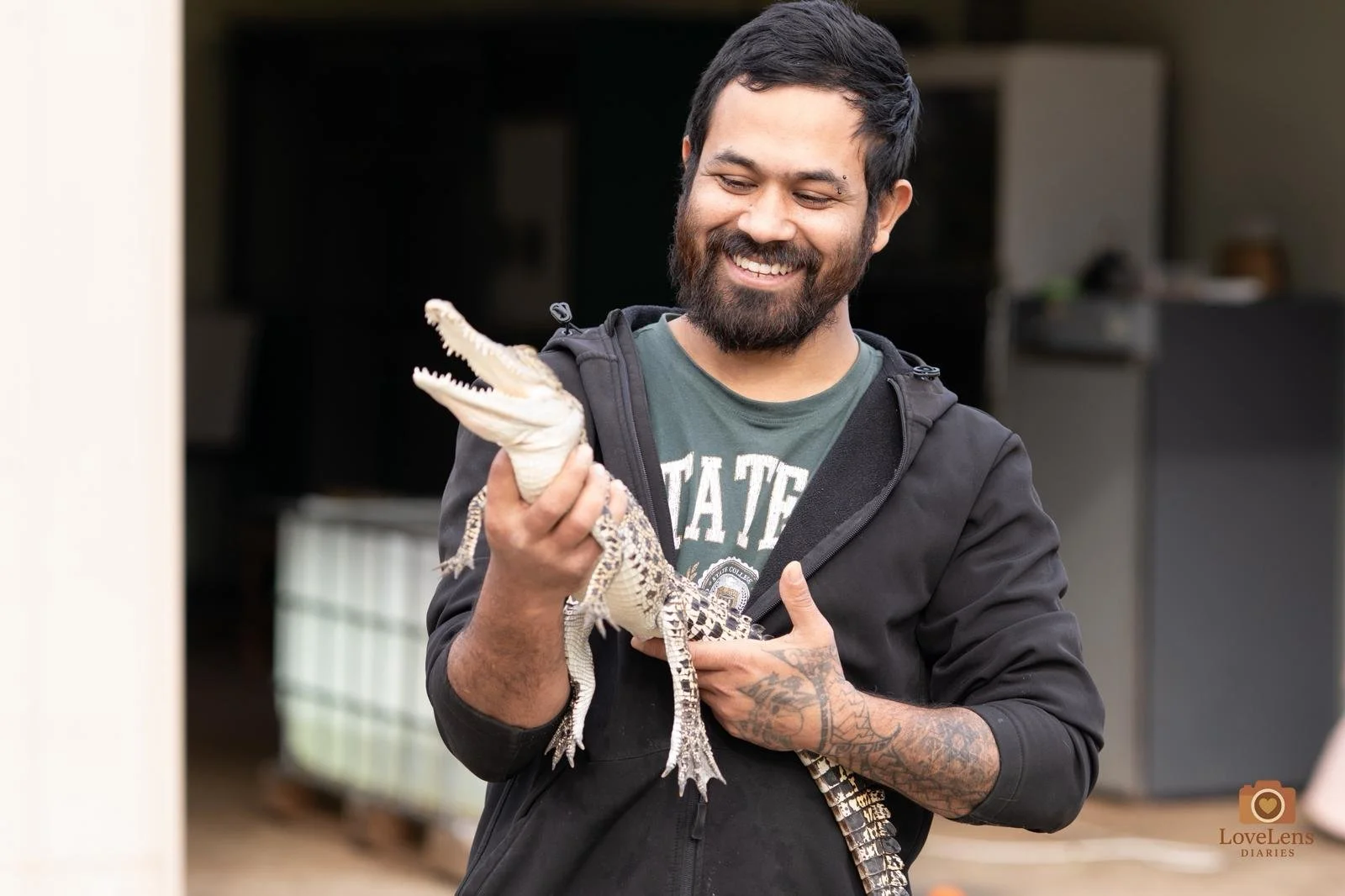 Man with tattoos and a beard smiling and holding a young crocodile indoors.