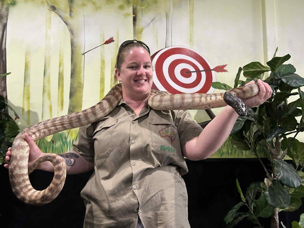 A woman in a zoo uniform holding a large snake draped around her shoulders, standing in front of a tan and green forest-themed backdrop with a red and white bullseye target and plants.