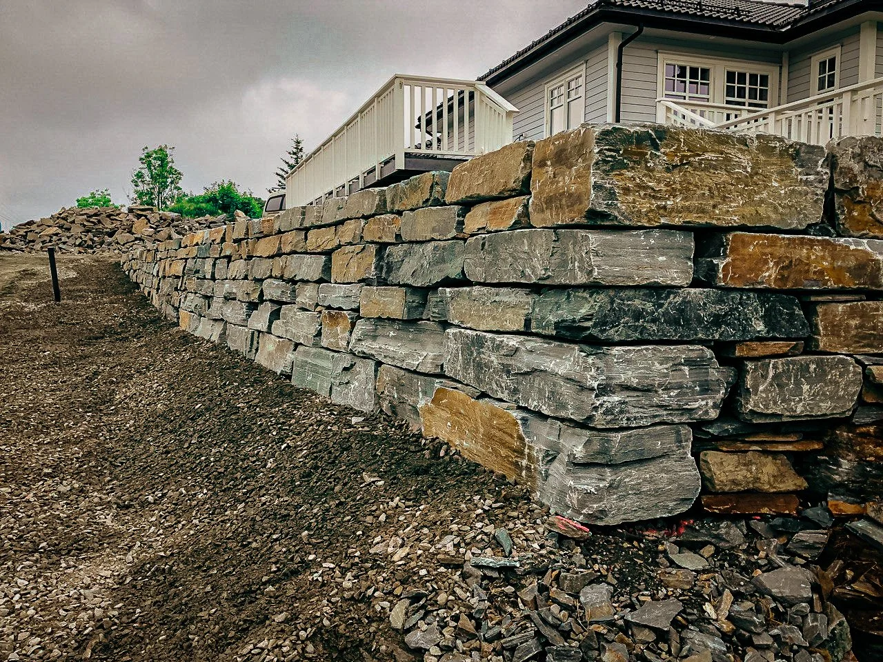 Et støttet mur av store steiner foran et hvitt hus med terrasse, med jord og småstein foran.