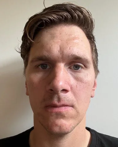 Nærbilde av en mann med kort, stritt hår og alvorlig uttrykk, mot en enkel, lys bakgrunn.