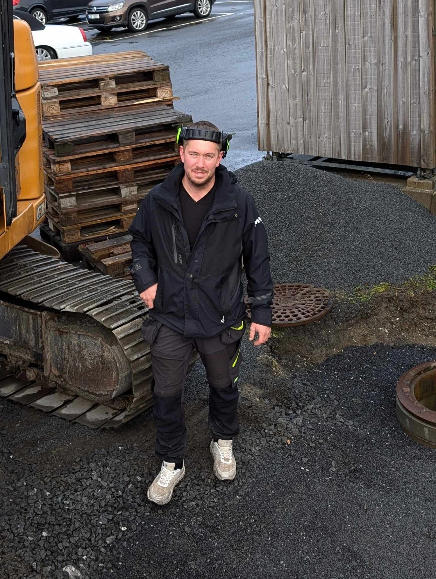 En mann står foran en gravemaskin og plass med nylagt asfalt, iført svart jakke, sorte bukser med gule detaljer og hvite sko, peker mot kamera.
