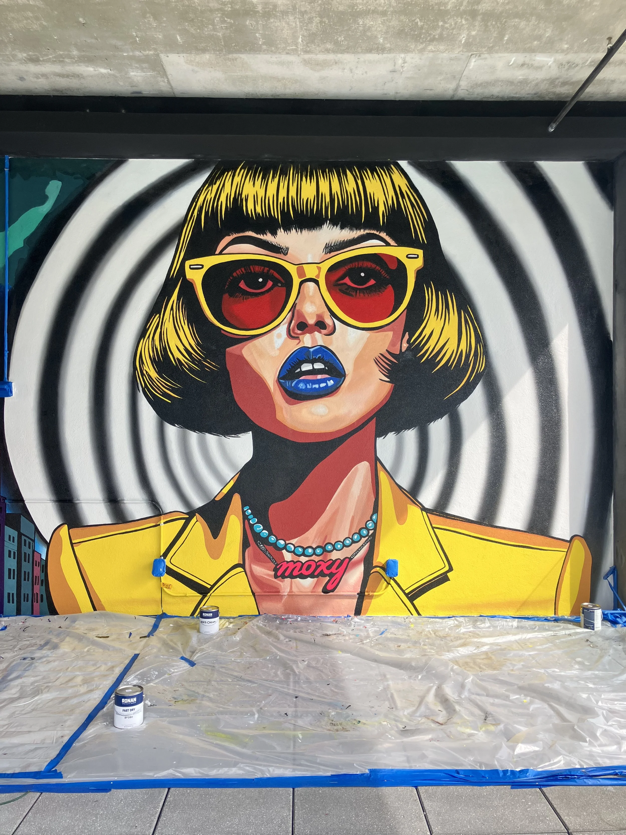 Colorful mural of a woman with yellow hair and sunglasses, with spiral black and white background. She is wearing a yellow jacket, a blue pearl necklace, and a pink 'moxy' necklace. There are paint cans and a plastic sheet in front of the mural.