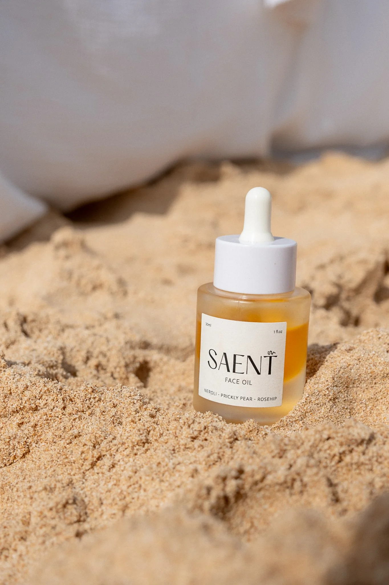 A small bottle of face oil labeled 'Saent' is placed on sandy ground, with a blurred white object in the background.
