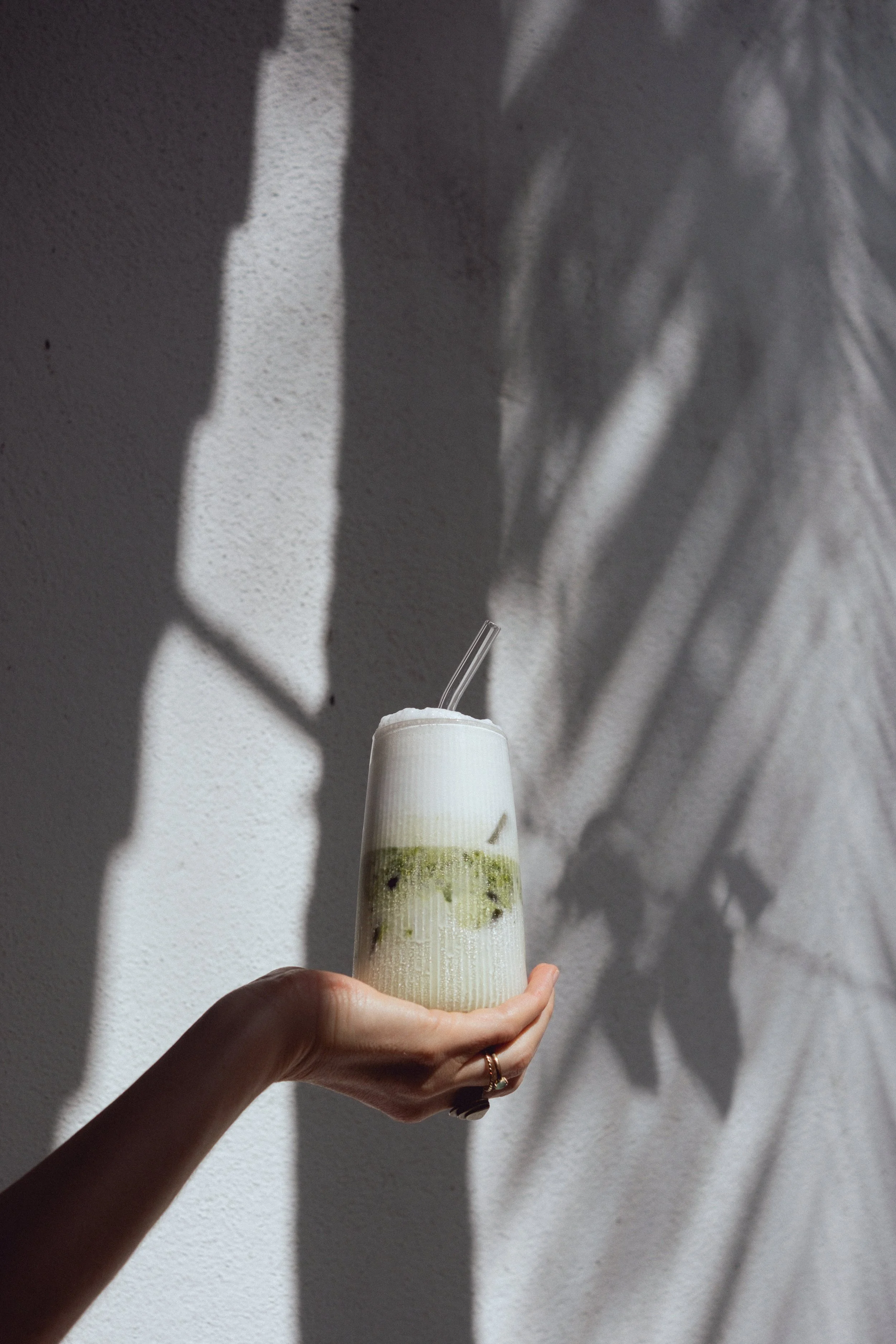 A person holding a tall, layered beverage with a straw, with shadows and light creating abstract patterns on the white wall behind.