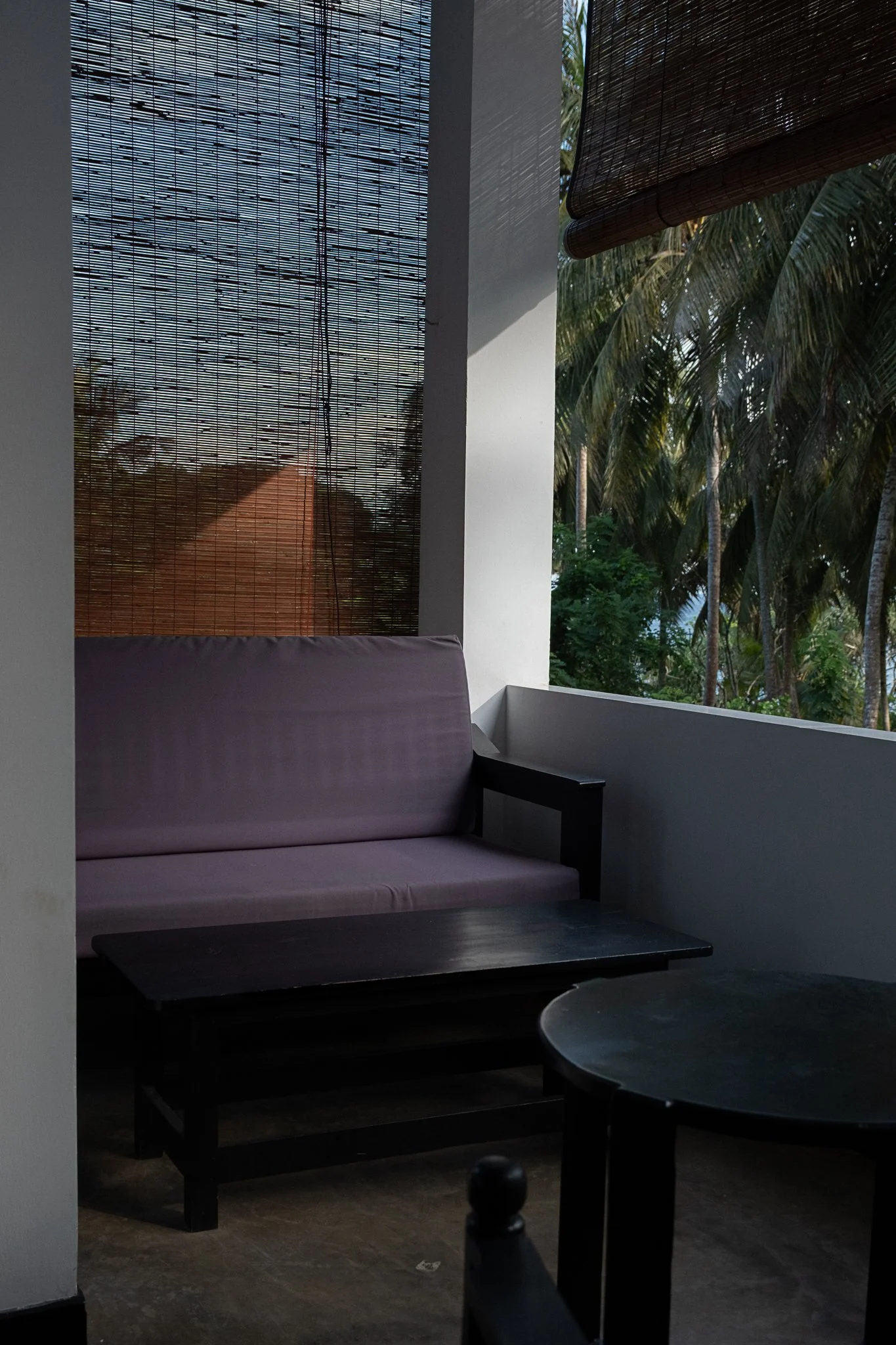 A cozy outdoor seating area with a cushioned bench, small tables, and a tropical background of palm trees visible through bamboo shades on a patio or balcony.