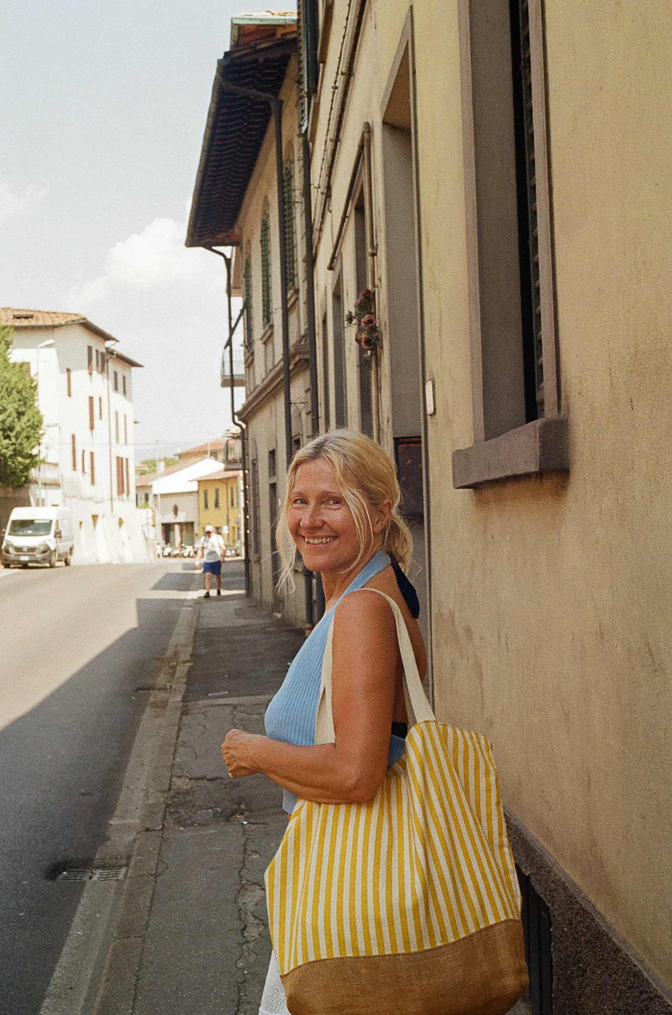 A smiling woman with blonde hair stands on a sidewalk in front of a beige building. She is wearing a light blue sleeveless top, a striped yellow and white bag, and brown shorts. There are other people and vehicles in the background on the street.