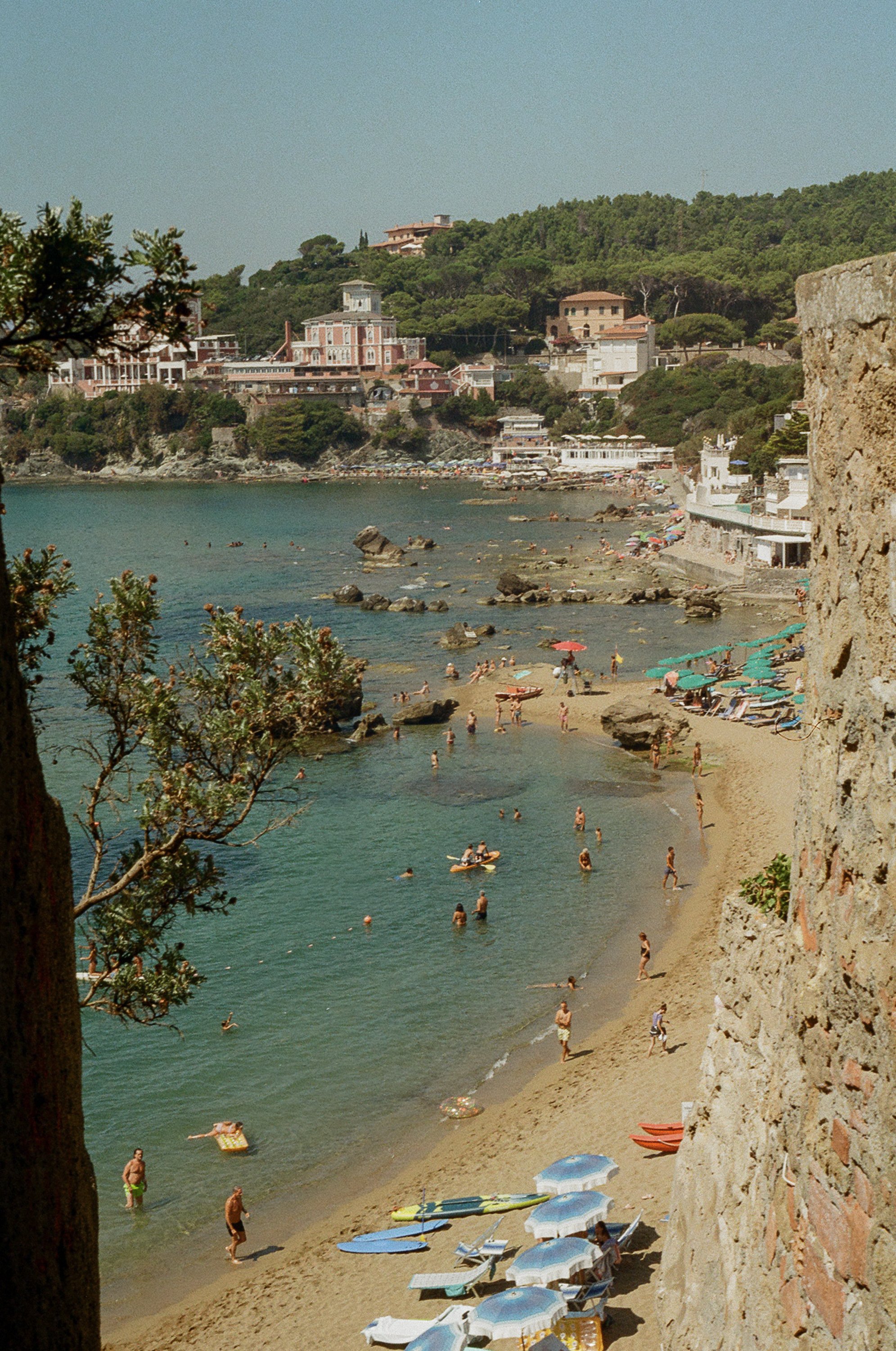 Beach scene with people swimming, sunbathing under umbrellas, and relaxing on chairs. The shoreline has rocks and a stone wall on the right. There are buildings and houses on a hillside in the background, with umbrellas and boats on the sand.