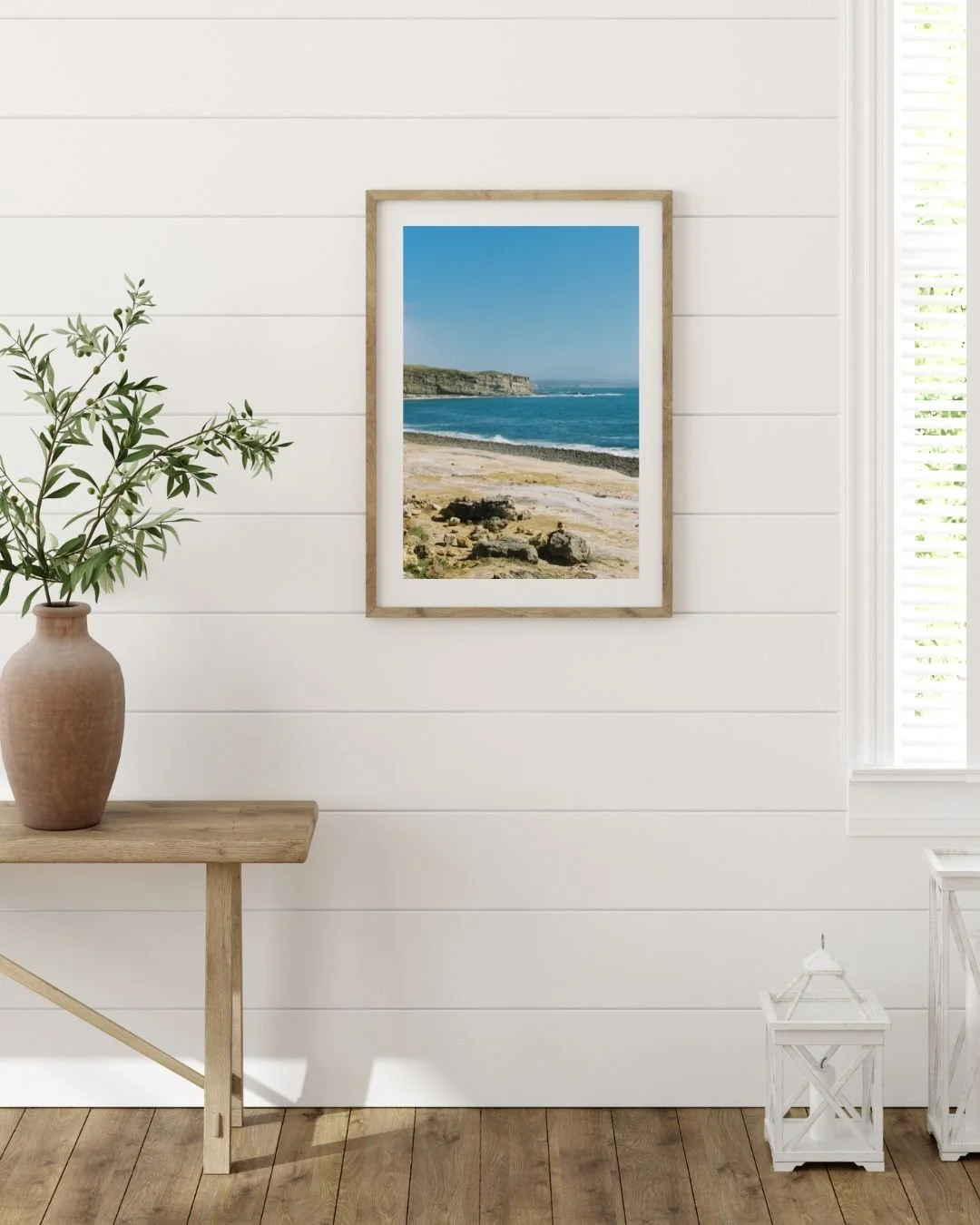 A framed picture of a beach scene with sand, rocks, ocean waves, and cliffs on a wall in a room with white shiplap walls, a wooden table with a plant in a clay pot, a white lantern, and a nearby window with blinds.
