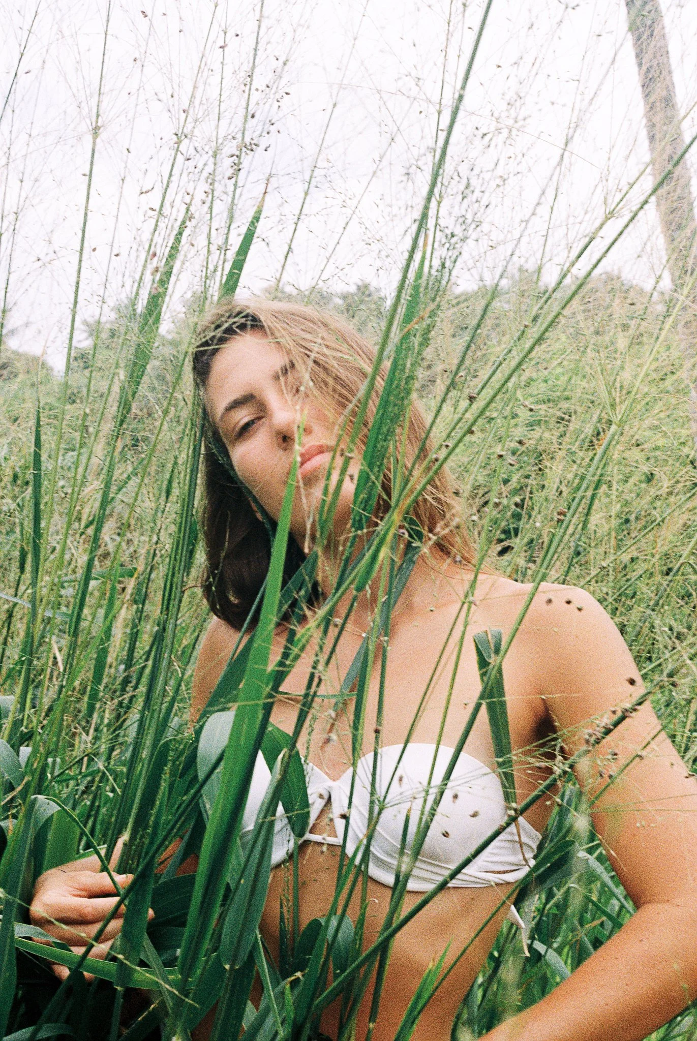 A woman with light skin and long brown hair sitting or lying among tall green grass or plants outdoors.
