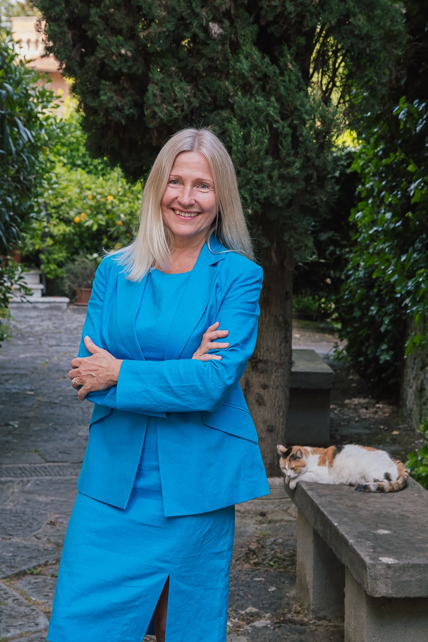 A woman in a blue suit standing outdoors with crossed arms, smiling, with a calico cat lying on a stone bench behind her, in a garden with green trees and bushes.