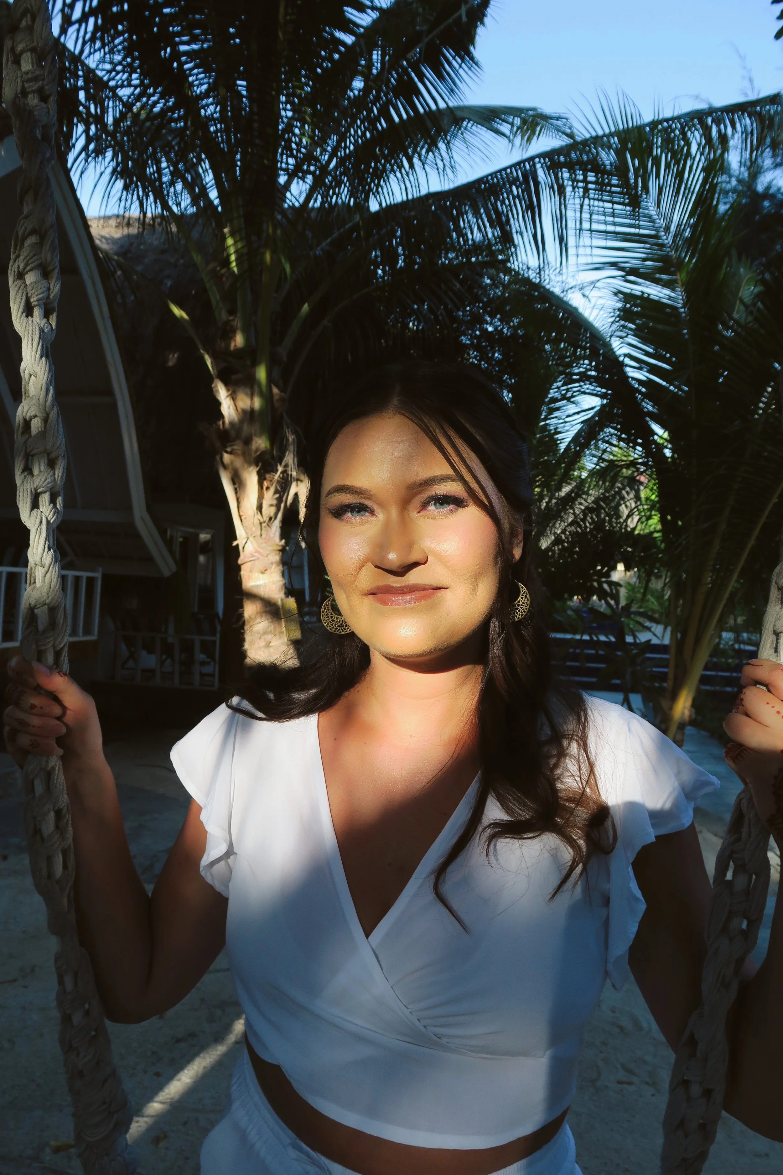 A woman with dark hair and fair skin smiling while holding onto ropes on a swing outdoors, with palm trees and a clear blue sky in the background.