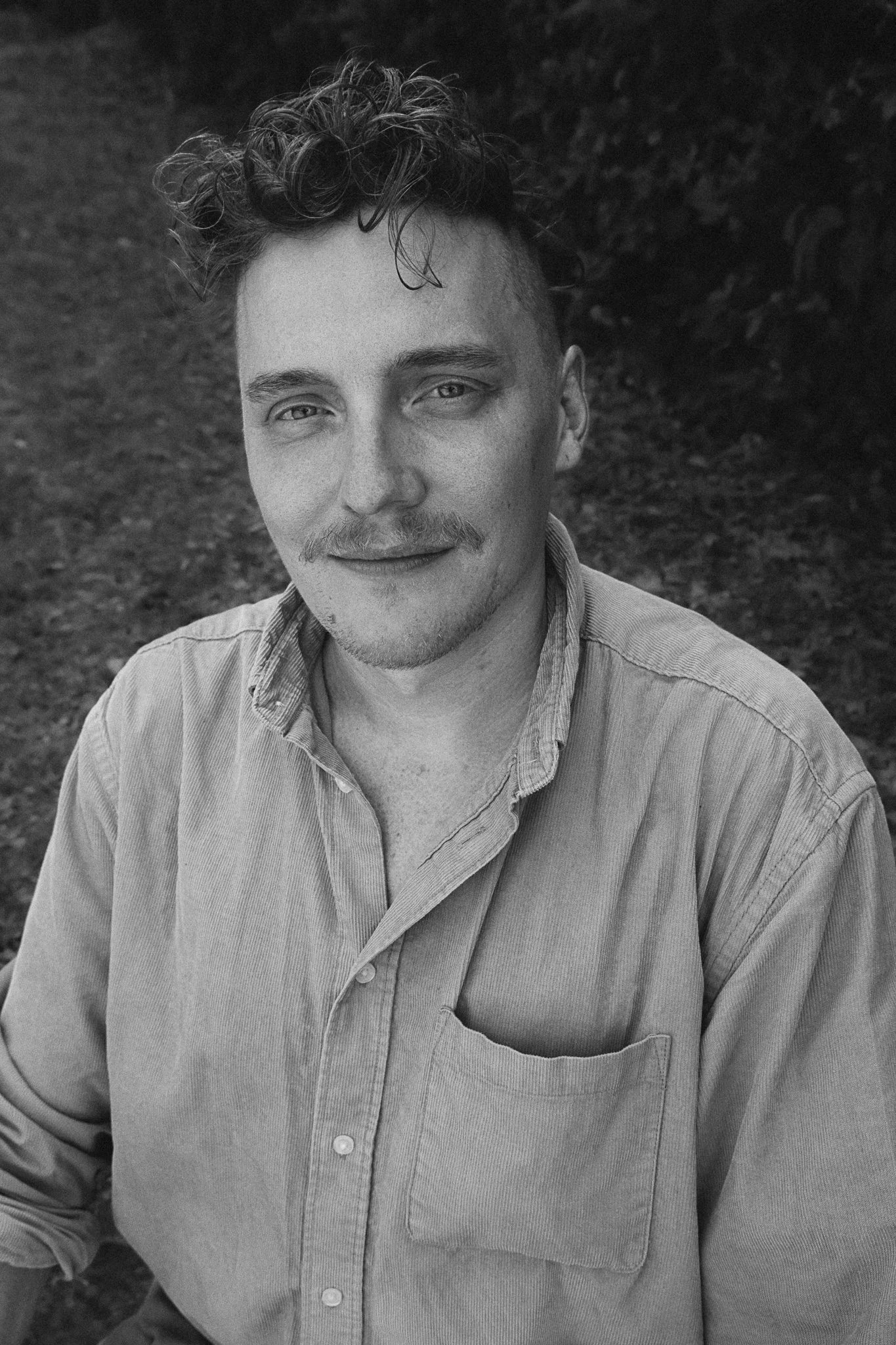 A black and white portrait of a young man with curly hair, light eyes, and a mustache, wearing a button-up shirt with a pocket on the chest.