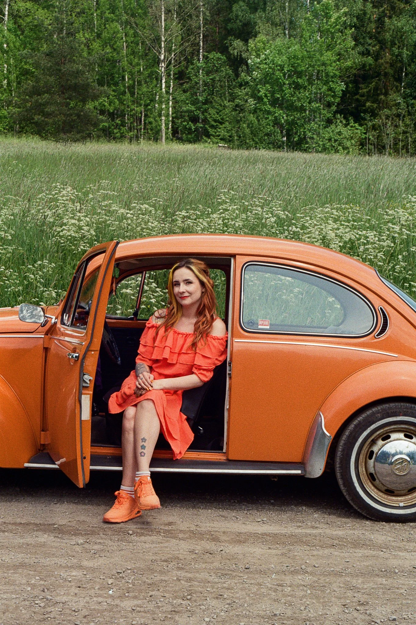 A woman with red hair, wearing an orange dress, orange shoes, and socks, sitting in the driver’s seat of an orange vintage car with the door open, on a dirt road with a lush green forest in the background.