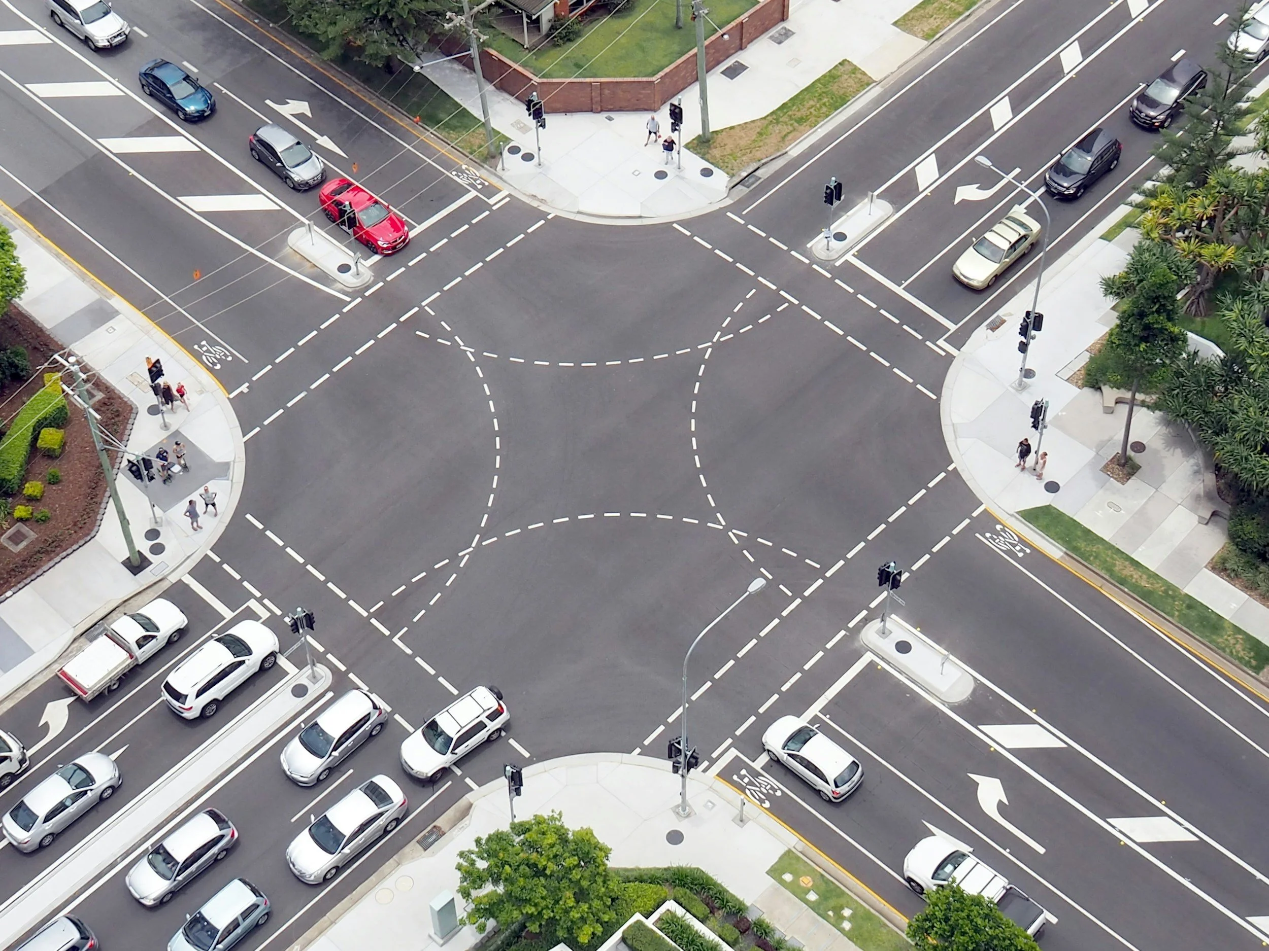 An aerial view of a city intersection with multi-lane roads, marked crosswalks, traffic lights, and several cars and pedestrians.