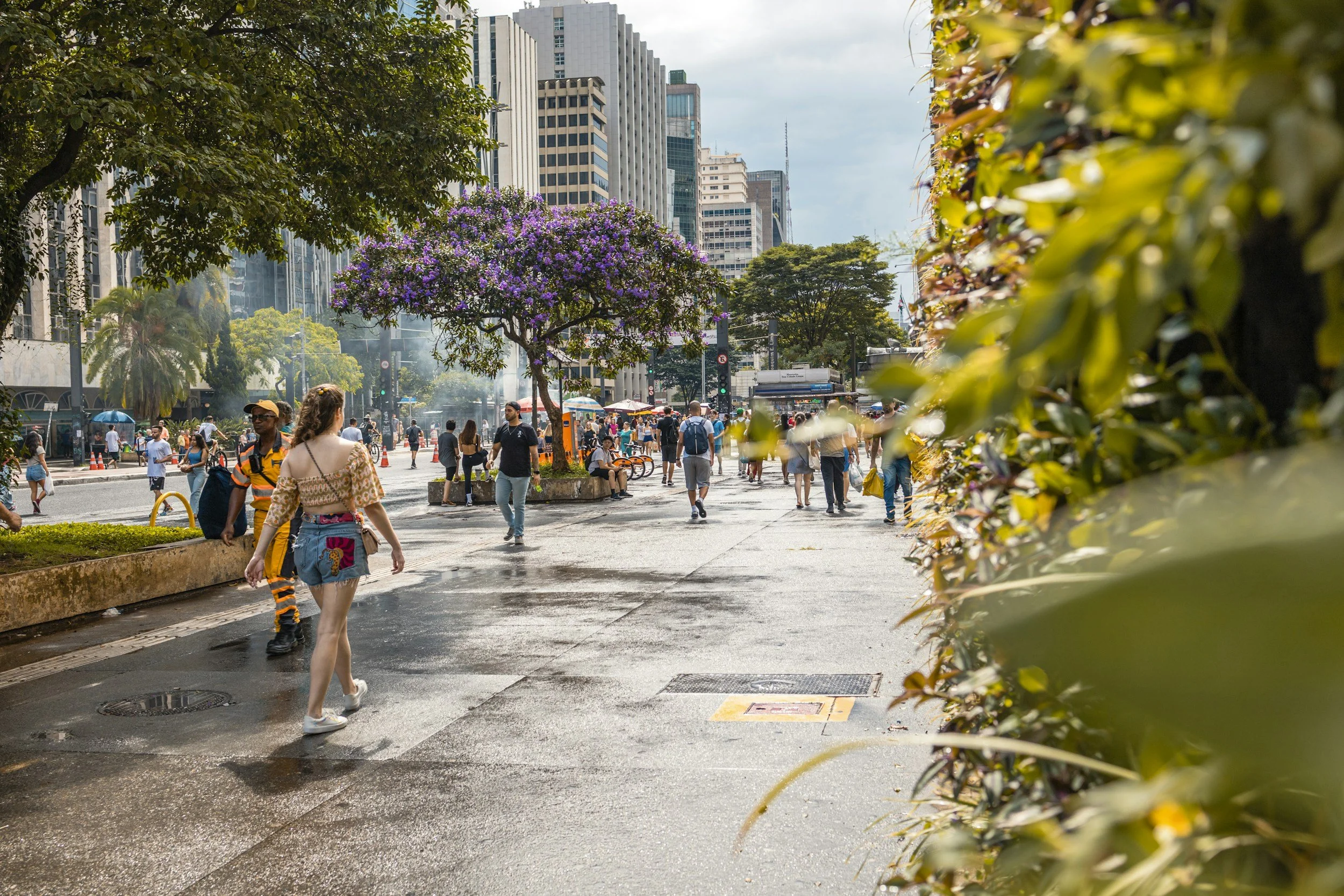 People walking in a city street with tall buildings and flowering trees, some people appear to be shopping or waiting at a bus stop, with a mix of sunny and cloudy weather.
