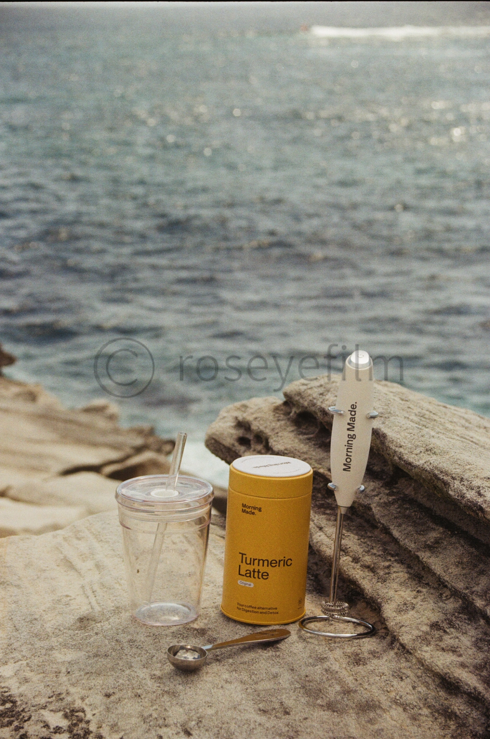 A turmeric latte in a yellow container, a clear plastic cup with a straw, a small silver spoon with salt, and a metallic sign that says 'Morning Made.' are arranged on a rock near the water at a beach.