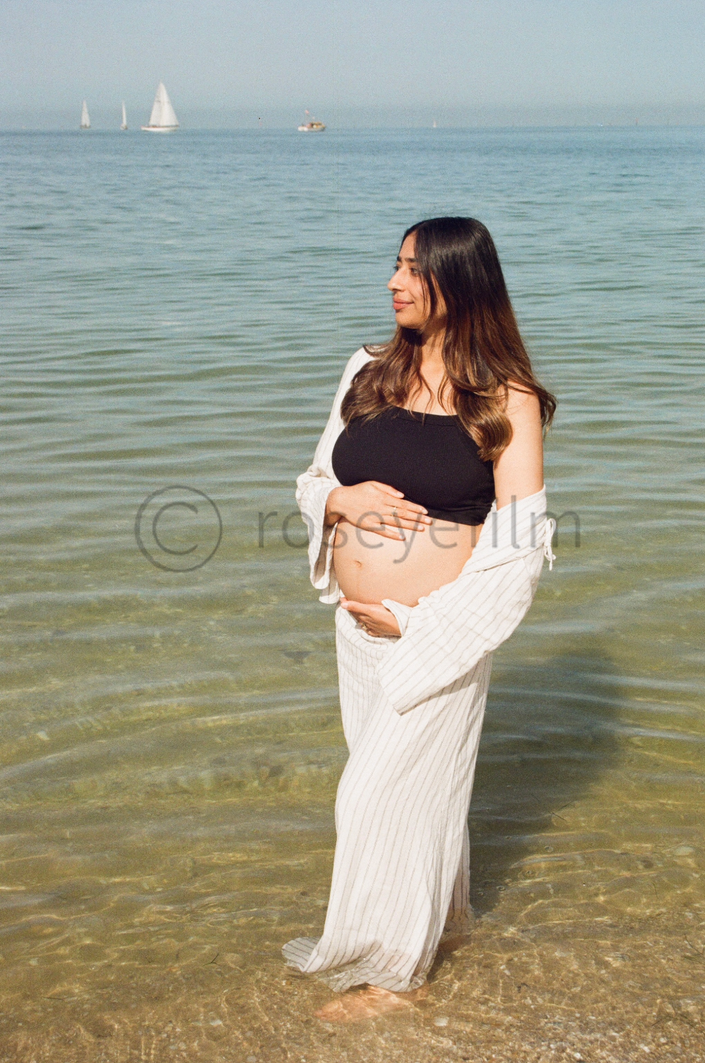 Pregnant woman standing in shallow water at the beach, with sailboats in the background.