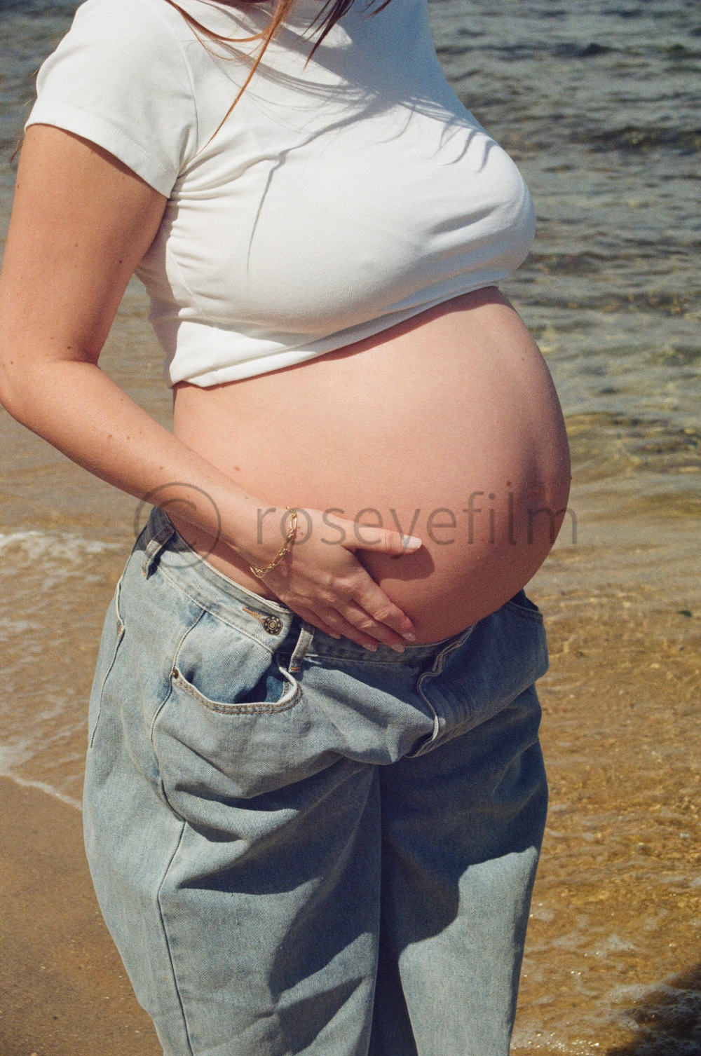 Close-up of a pregnant woman standing on the beach, wearing a white t-shirt and jeans, with her hand on her belly.