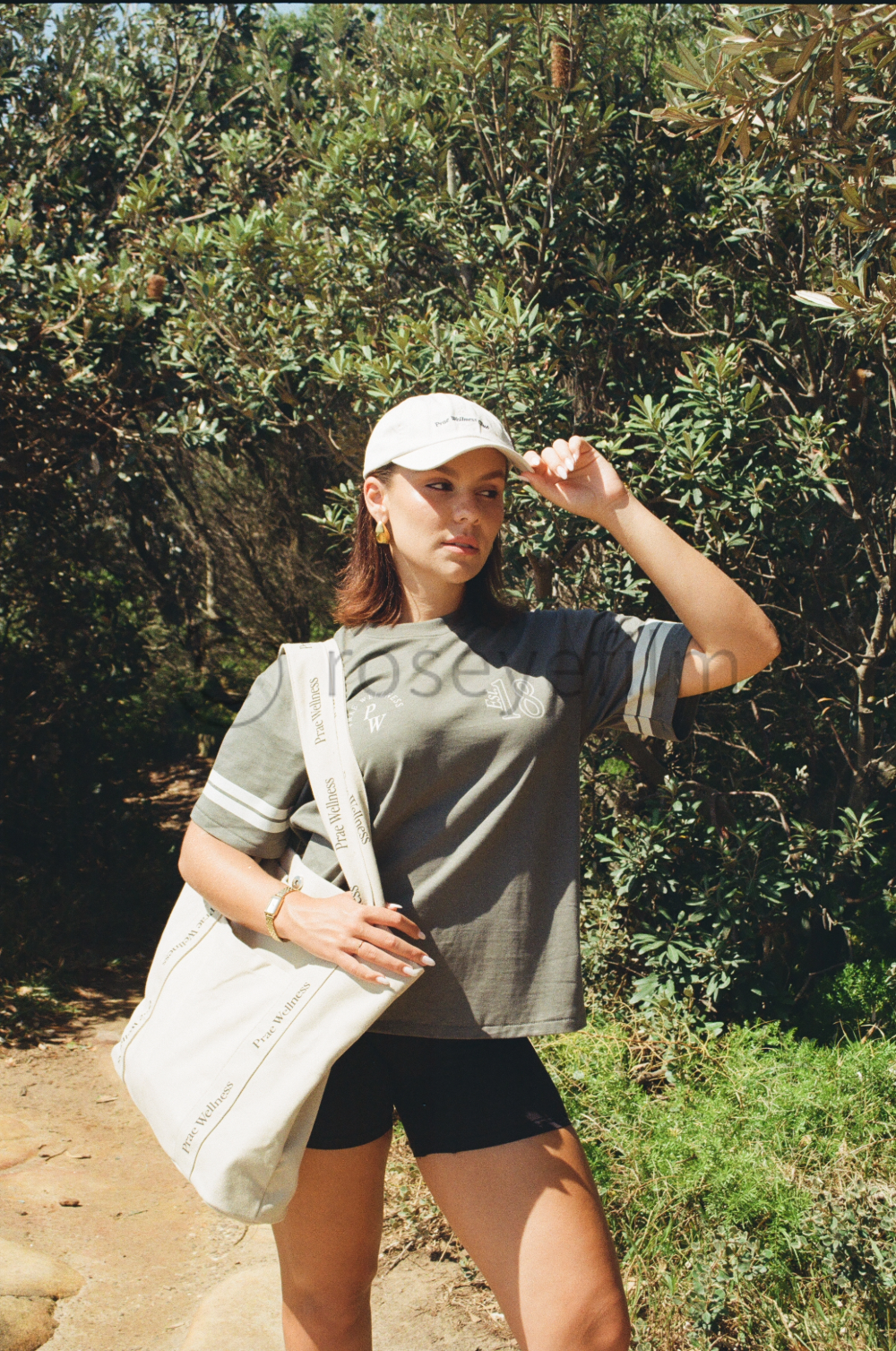 A young woman in a grey t-shirt, black shorts, a white baseball cap, and gold earrings standing outdoors on a dirt path surrounded by green foliage, holding a large white tote bag.