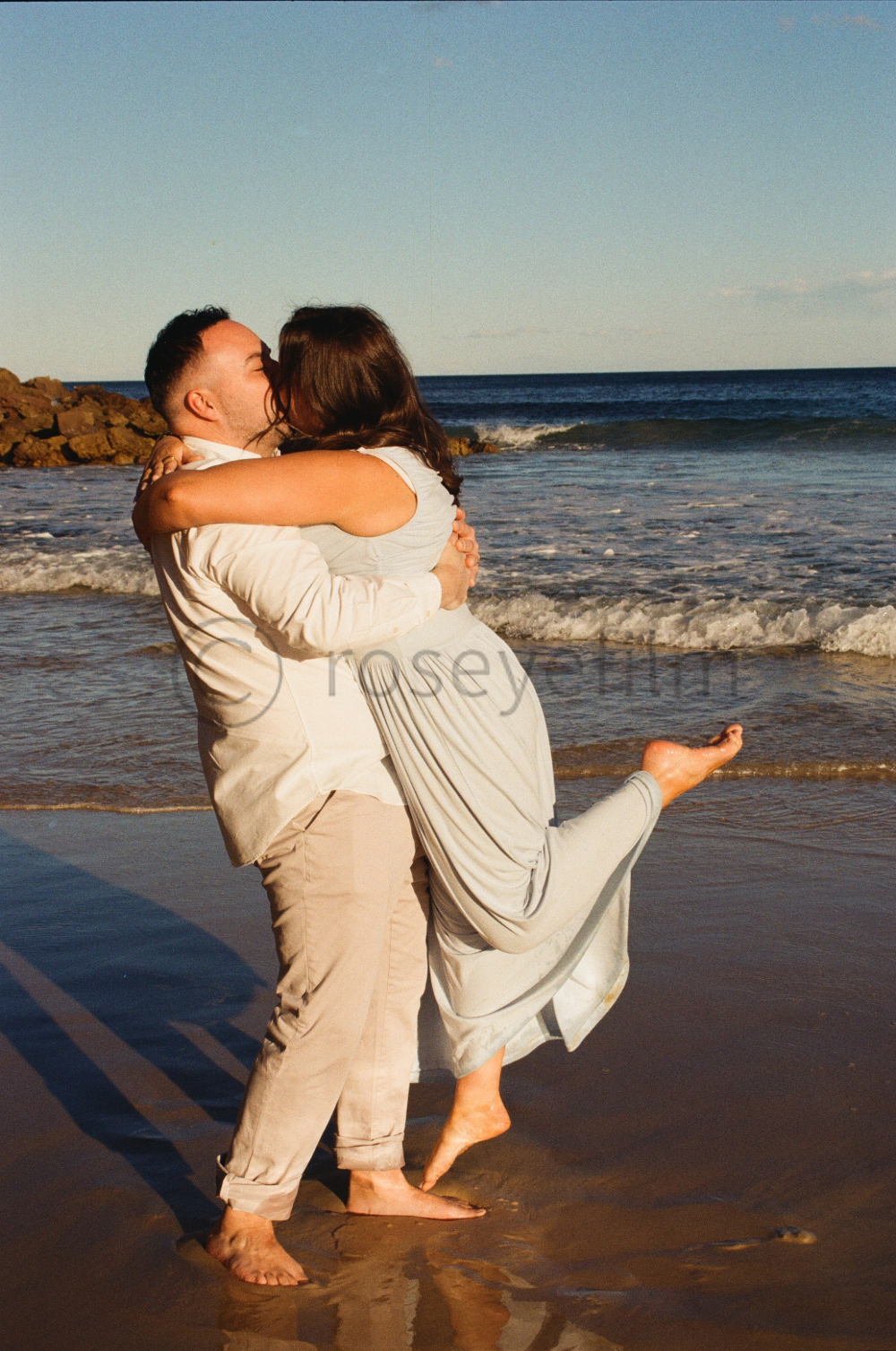 A couple hugging and kissing on the beach during sunset, with ocean waves in the background.