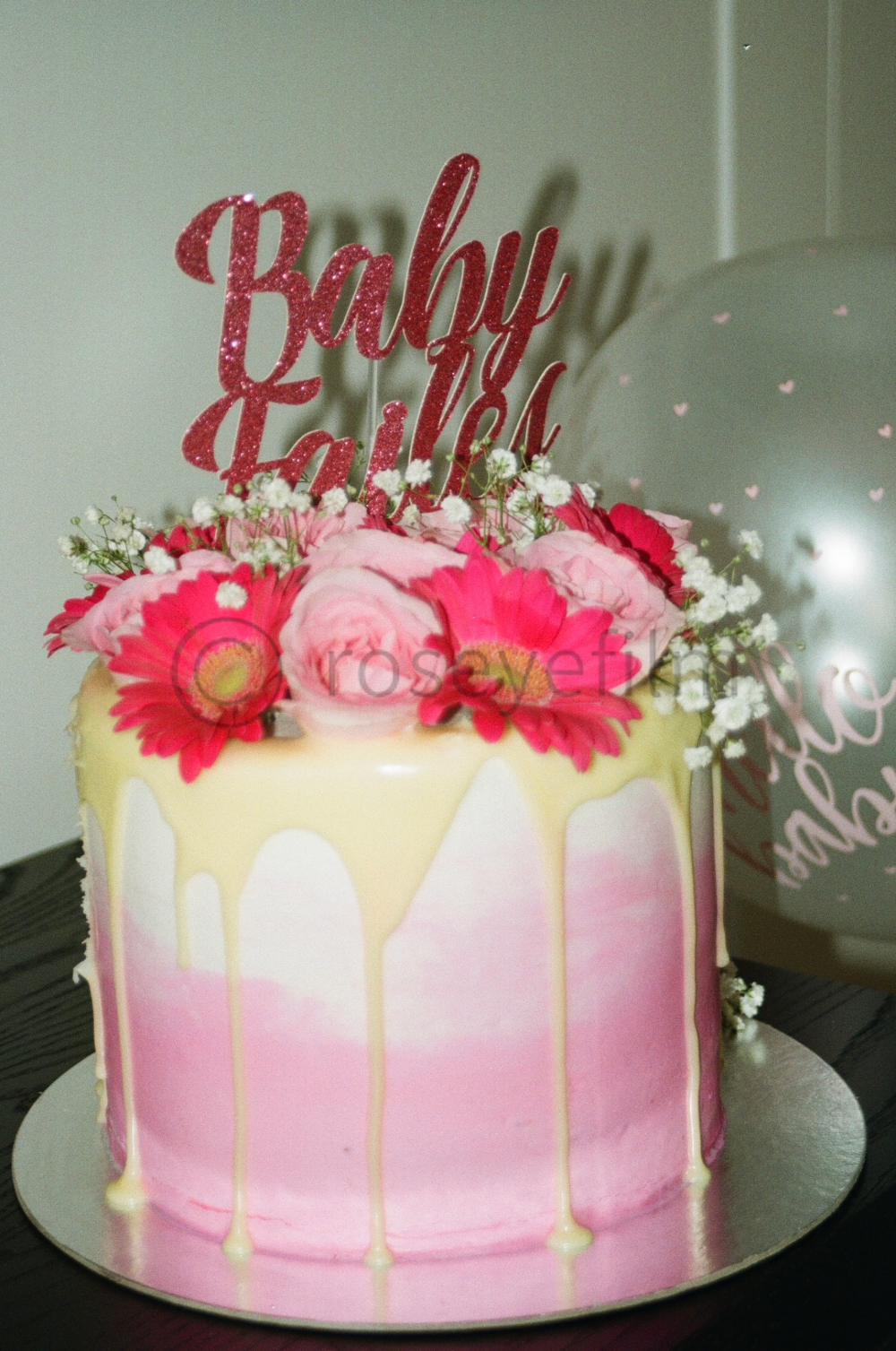 A pink and white drip cake decorated with pink and red flowers and a glittery red "Baby" cake topper for a celebration.
