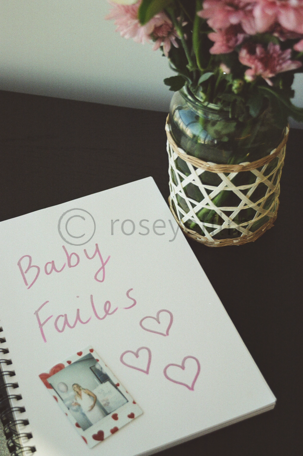 A notebook with the words 'Baby Fail' written on it, decorated with hearts and a small photo of a woman, placed next to a glass vase with pink flowers inside.