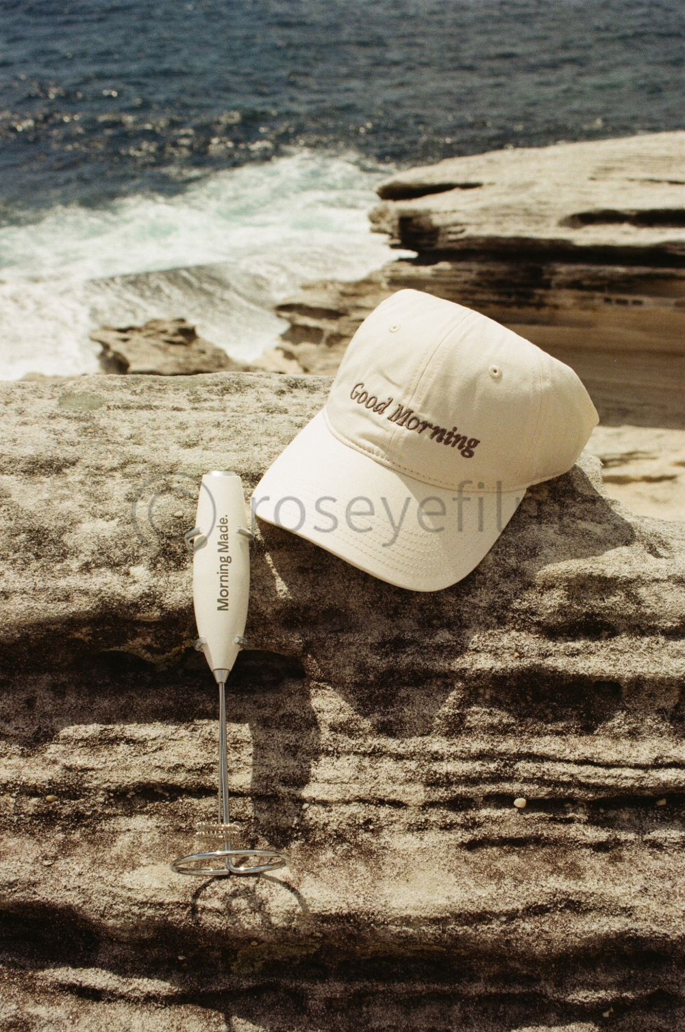 A white baseball cap embroidered with 'Good Morning,' a small white flashlight with 'Morning Made.' printed on it, placed on a rocky surface near the ocean with waves and cliffs in the background.