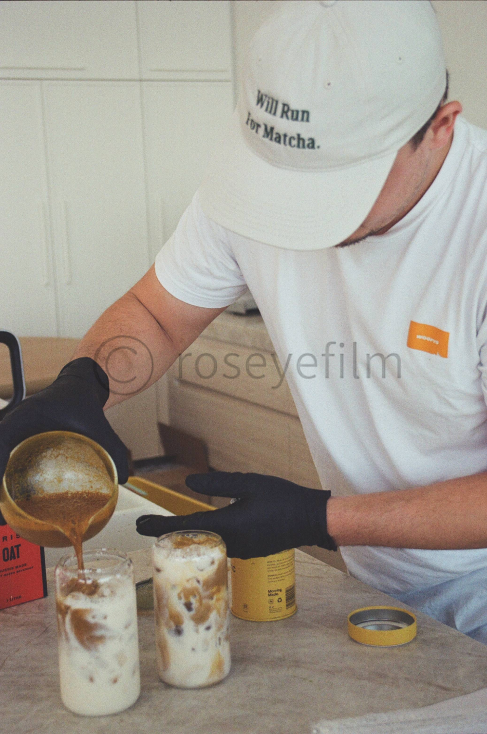 A person wearing a white shirt, black gloves, and a white cap labeled 'Will Run for Matcha,' pours a brown liquid into two glass jars filled with ice and milk.