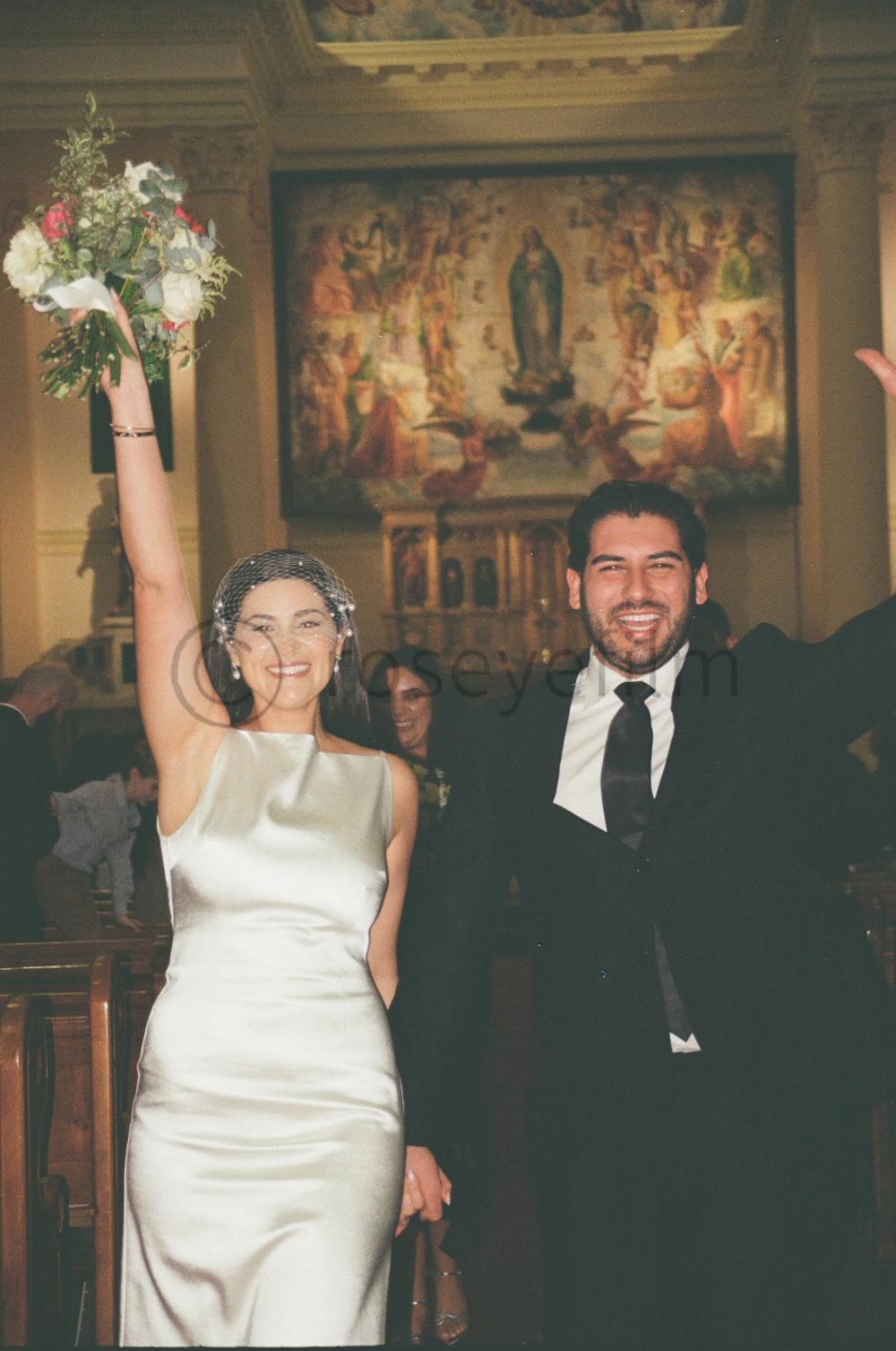 A bride and groom celebrating their wedding inside a church, with the bride holding a bouquet and raising her arm, and the groom smiling with his arm raised.