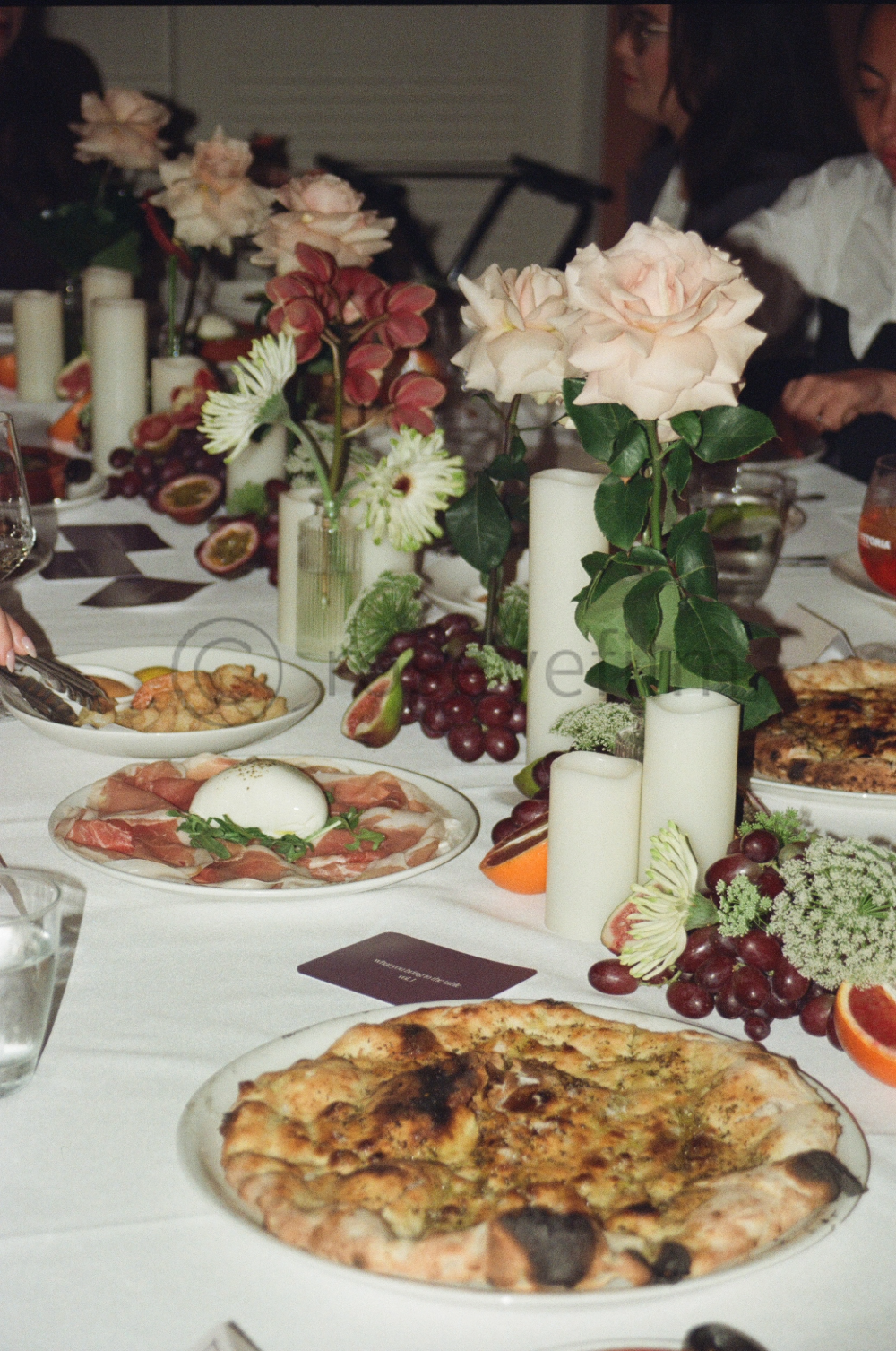 A festive dinner table decorated with pink and white roses, candles, and assorted fruits, including grapes and halved passionfruits. The table has pizza, a plate with prosciutto and a poached egg, and glasses of drinks, suggesting a social celebratio