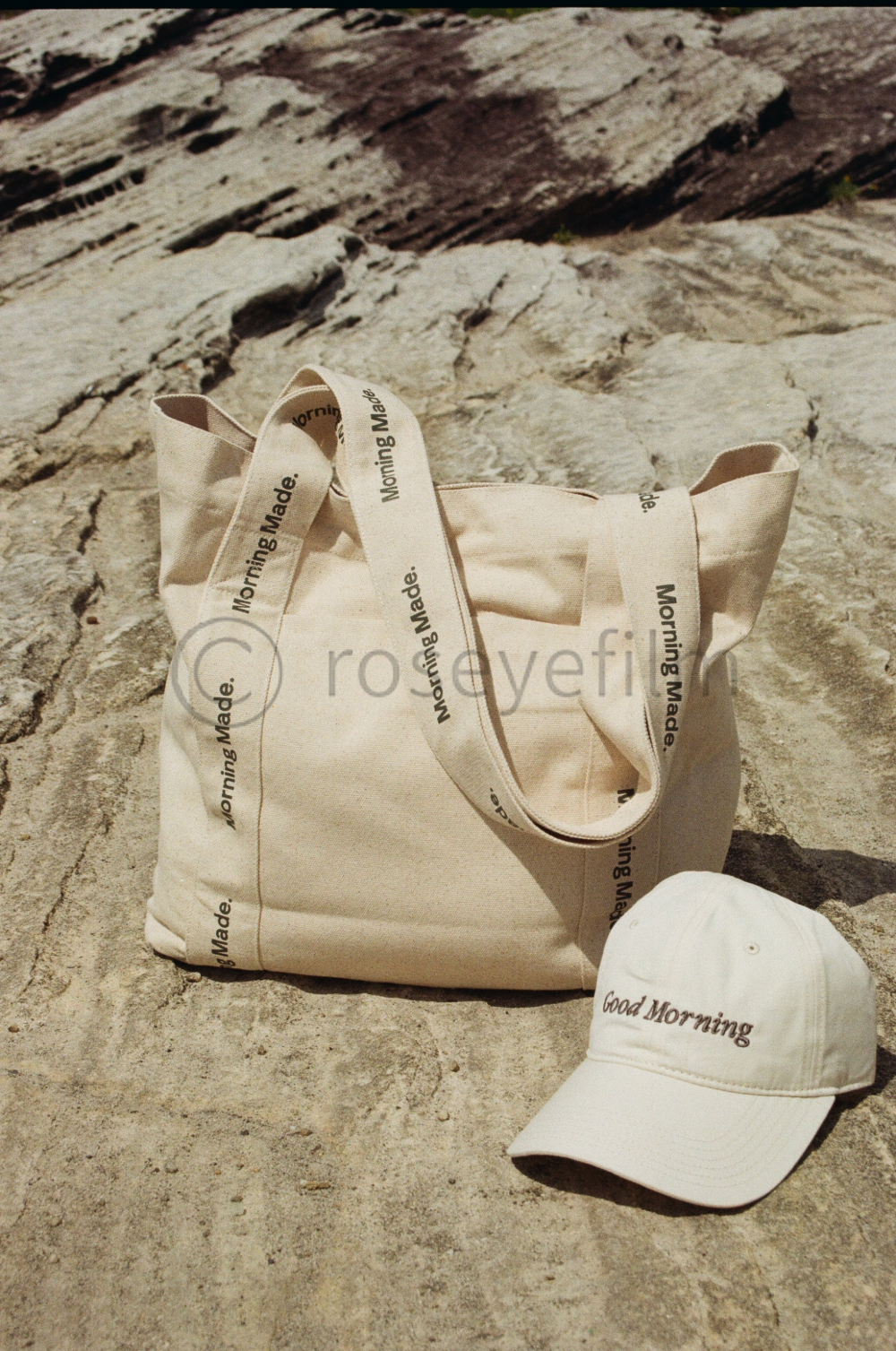 A beige tote bag and a beige baseball cap on rocky ground outdoors. The tote bag has black text on the straps that read "Morning Made." The baseball cap has black text that says "Good Morning." The background appears to be a rough rock surface.