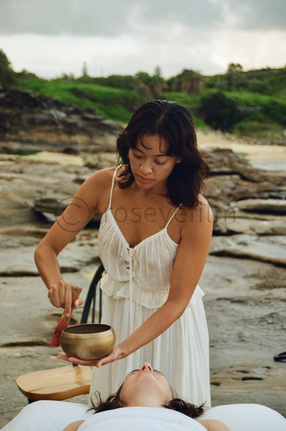 A woman giving a sound healing or meditation treatment outdoors near a rocky shoreline with green hills in the background, during cloudy weather.